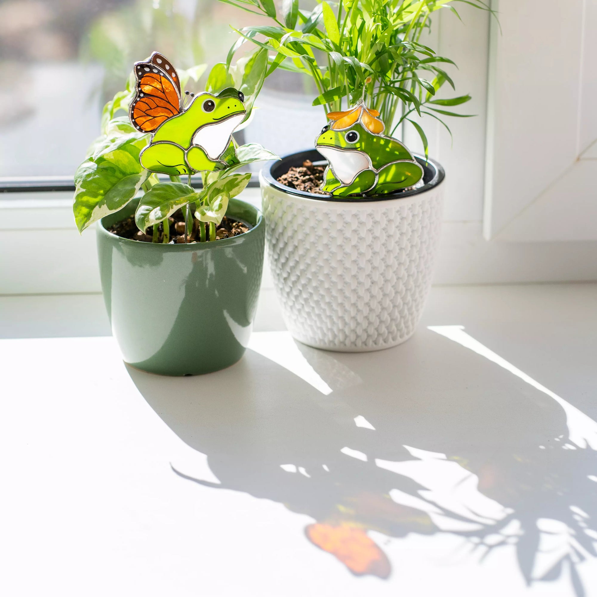 Stained glass plant stakes with decorative frogs and butterflies on a windowsill.