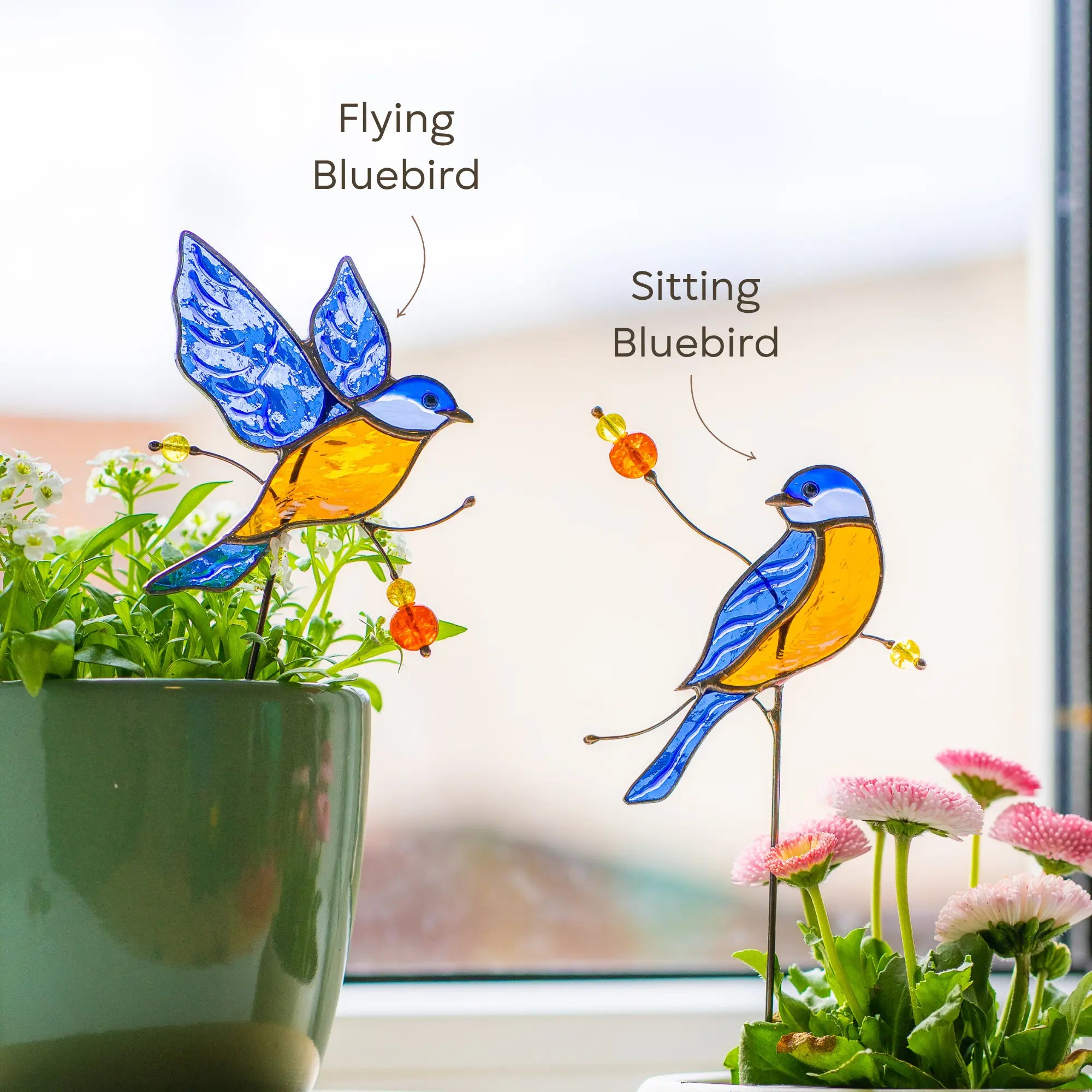 Two stained glass bluebird plant stakes, one flying and one sitting, in front of a window with flowers.