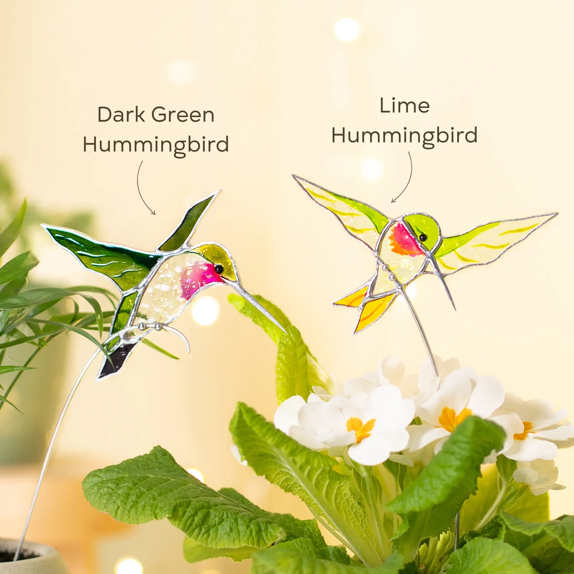 Variation of two stained glass hummingbird plant stakes, one dark green and one lime colored, on a plant with flowers.