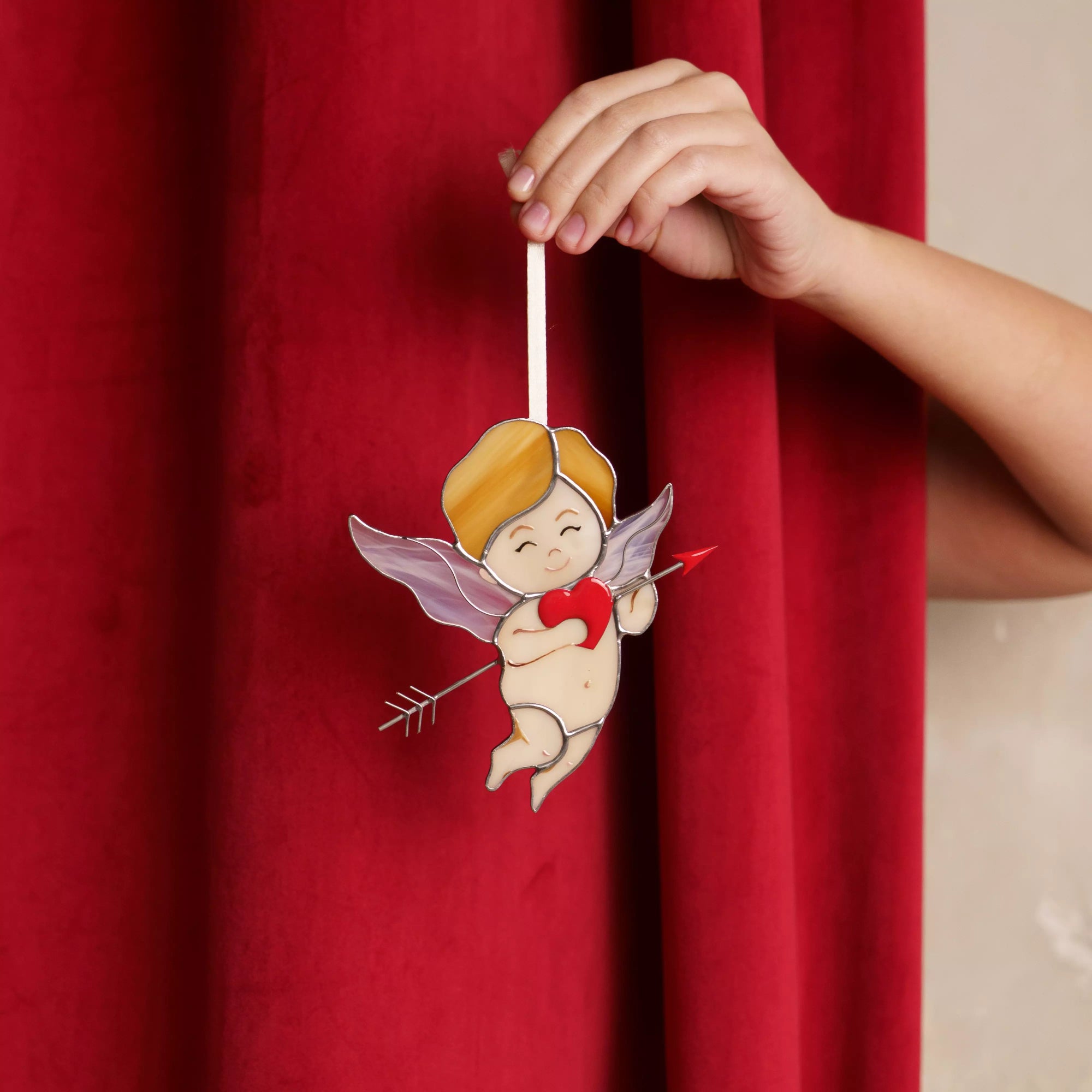 Stained glass cupid suncatcher held against a rich red curtain background, highlighting the red heart and arrow.