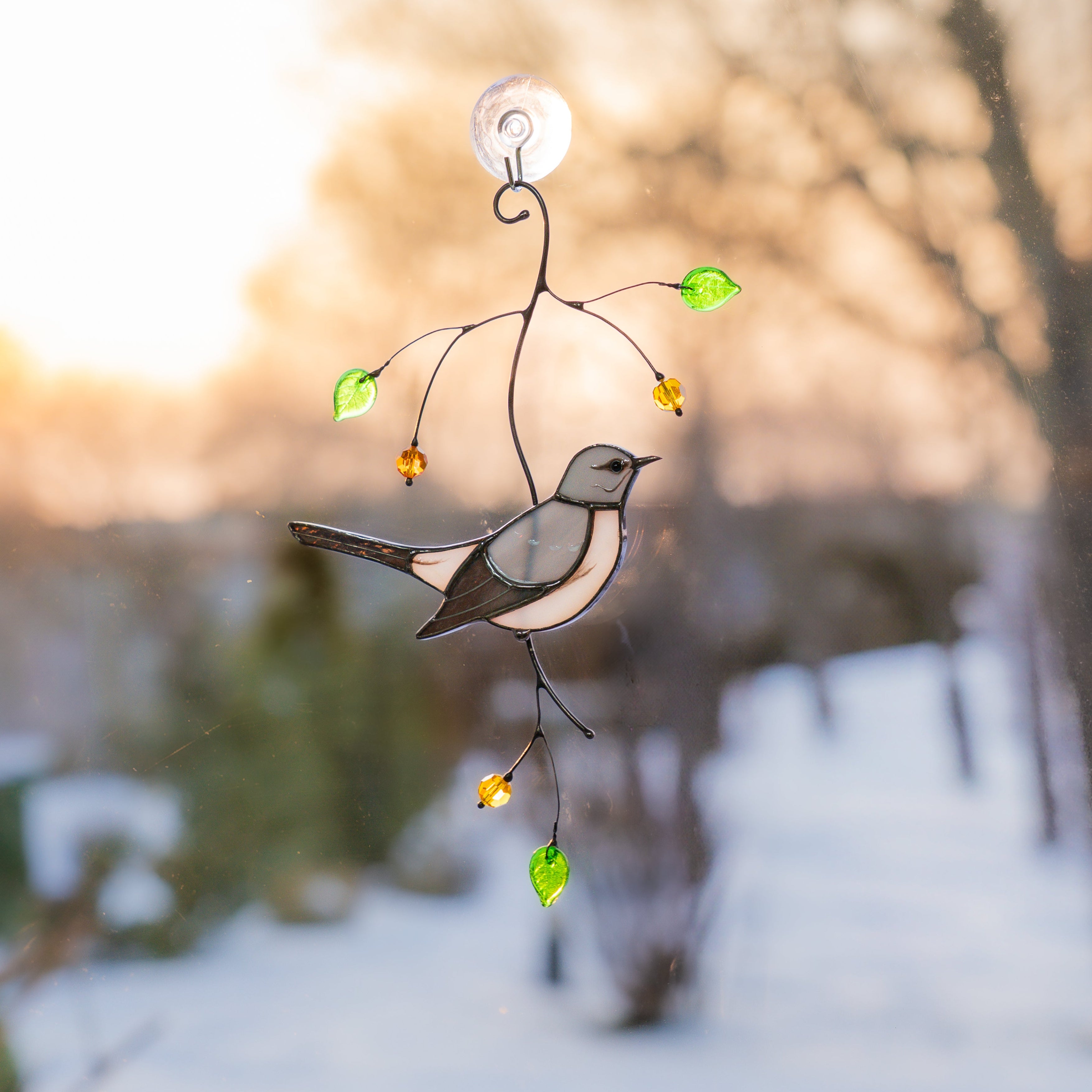 Stained glass mockingbird with green glass leaves hanging against evening winter sunset.