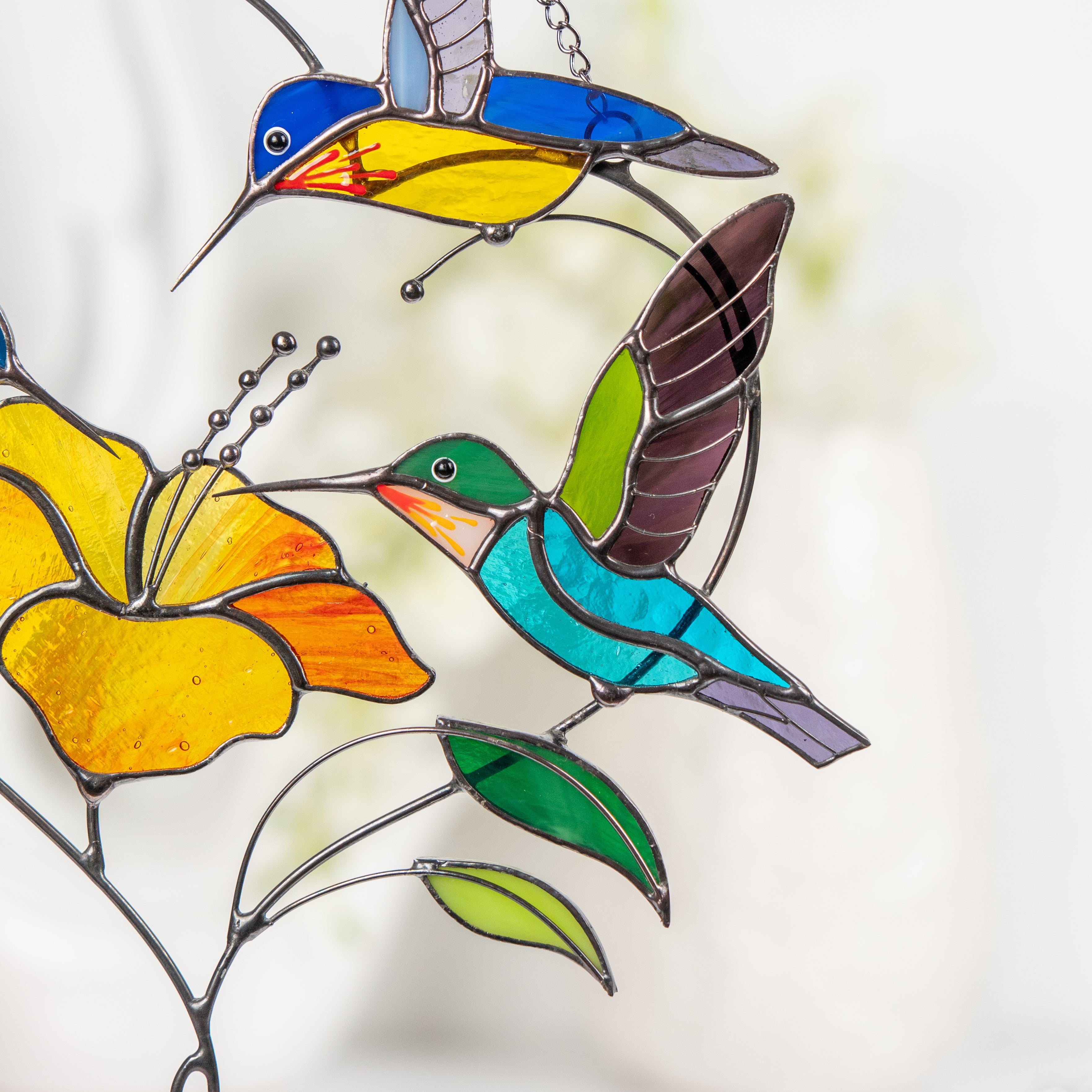 Two stained glass hummingbirds in turquoise and yellow hover around a hibiscus flower.
