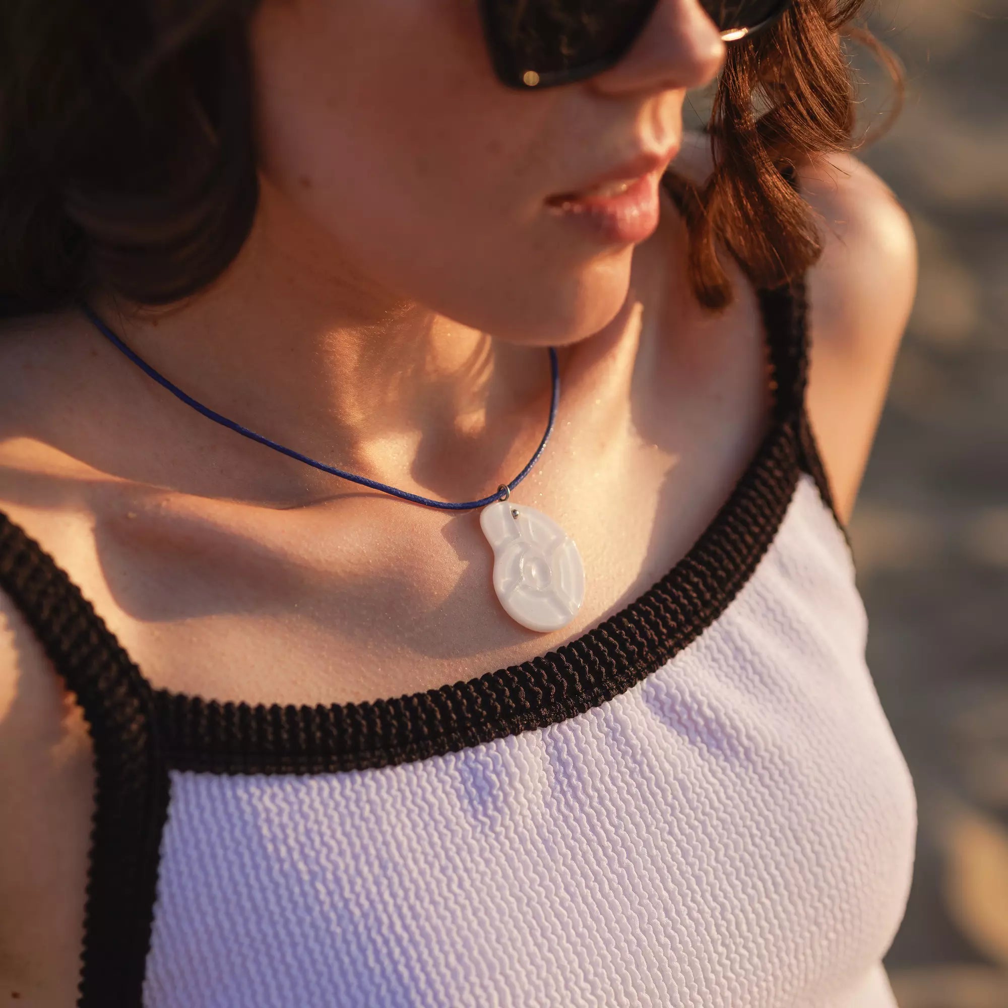 Close-up of woman wearing a white stained glass nautilus pendant on a dark blue cord, with sunlight highlighting her collarbone.