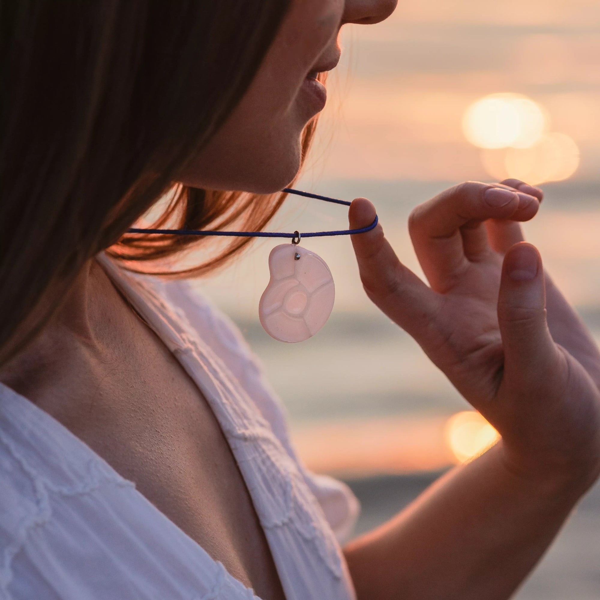 Woman holding a white nautilus-shaped stained glass pendant on a blue cord, with soft sunset light in the background.