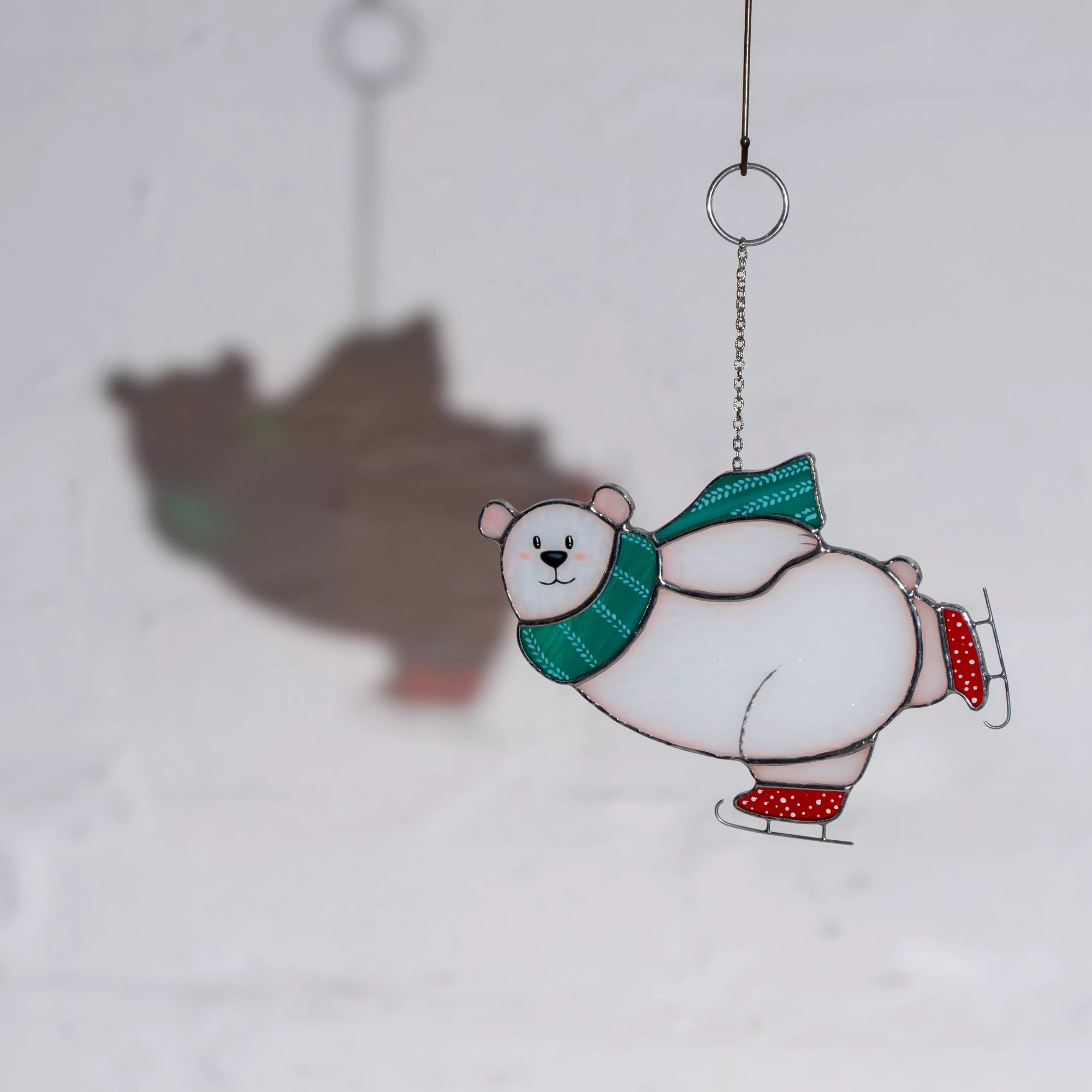 Stained glass window hanging ornament of a polar bear with a green scarf and red boots, casting a soft shadow on a white background.