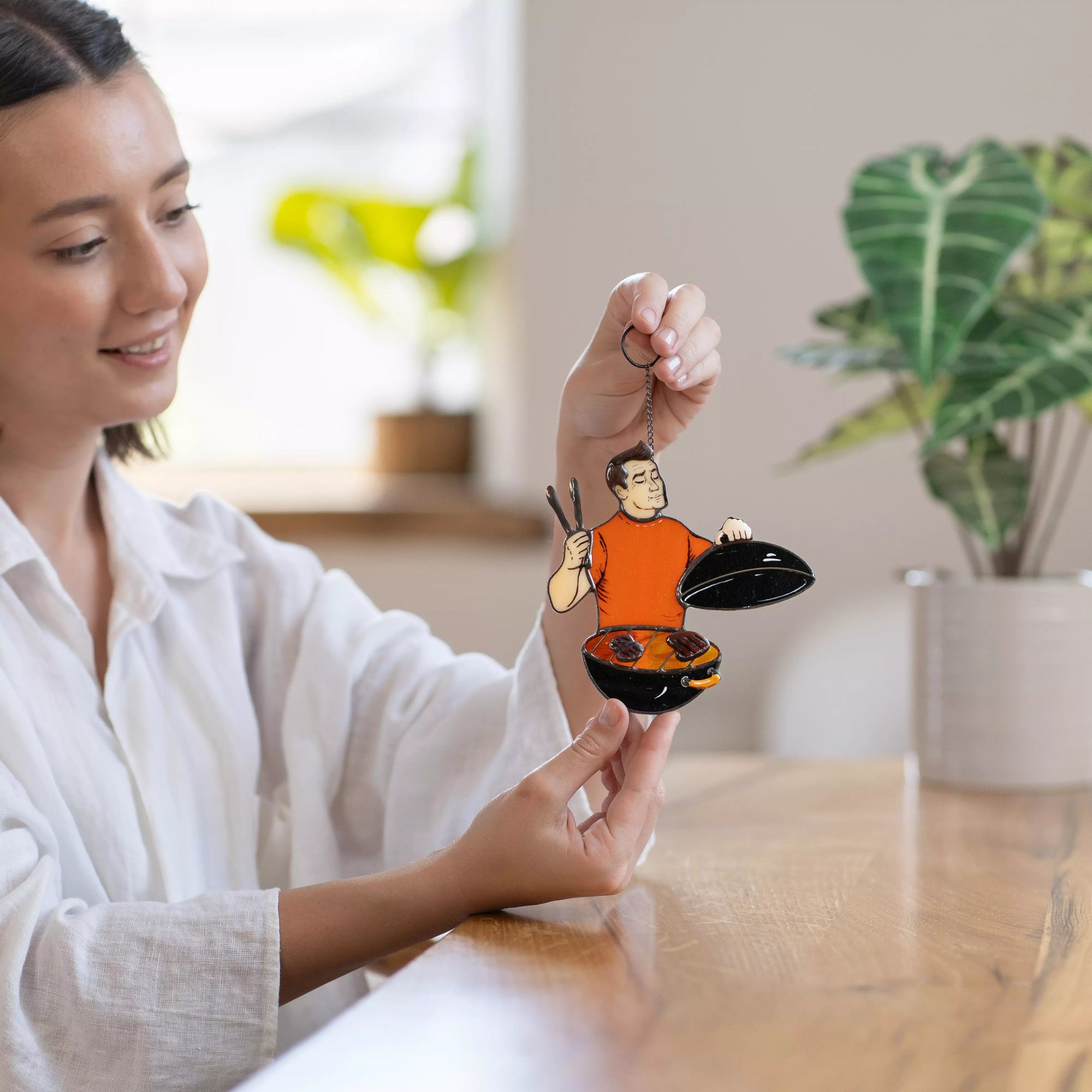 Woman holding stained glass ornament with an illustration of a person cooking, in a home setting.
