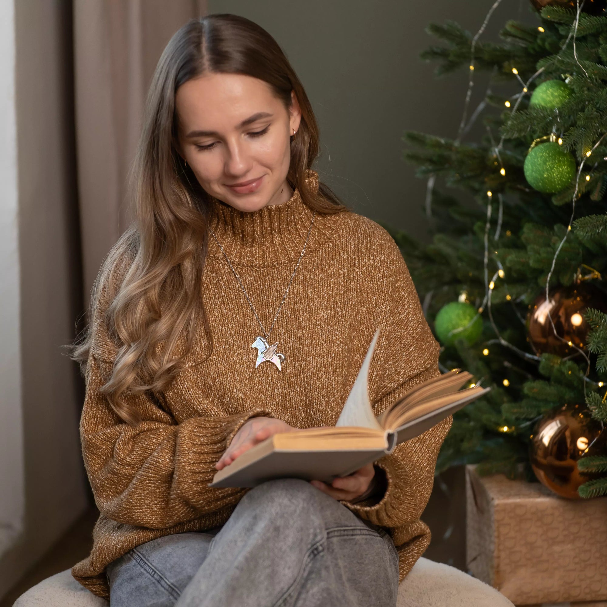 Woman with a stained glass pegasus necklace on the neck reading a book in front of a decorated Christmas tree.