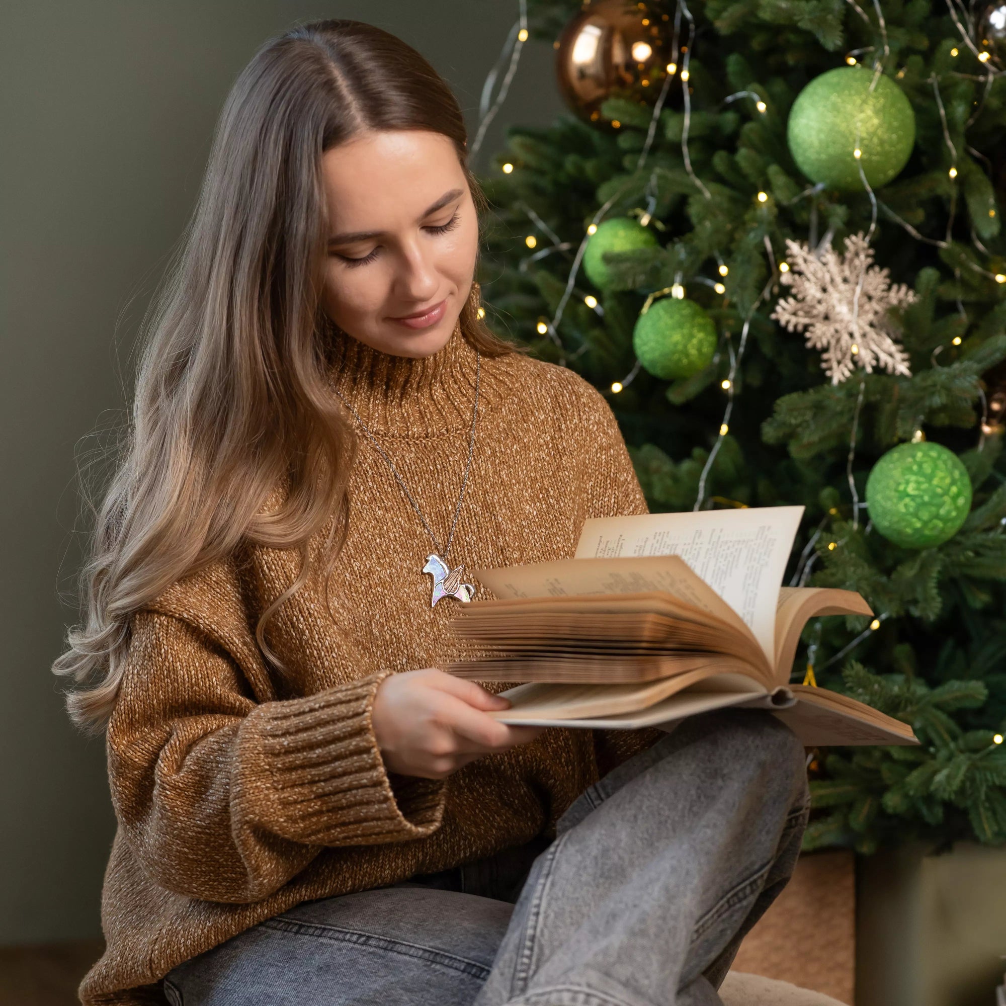 Woman with pegasus necklace on her neck reading a book in front of a decorated Christmas tree