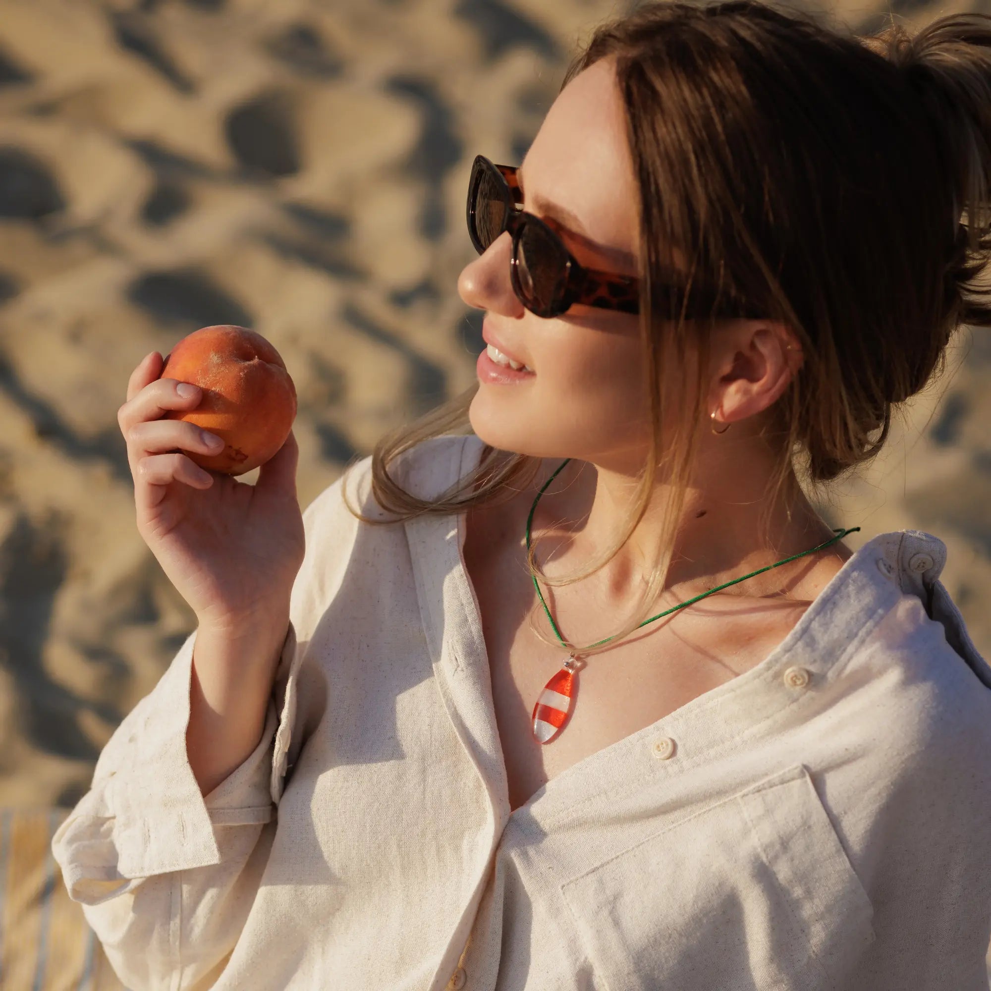 Smiling woman in sunglasses and linen shirt holding a peach, wearing Coral fish fused pendant necklace with green cord on the beach.