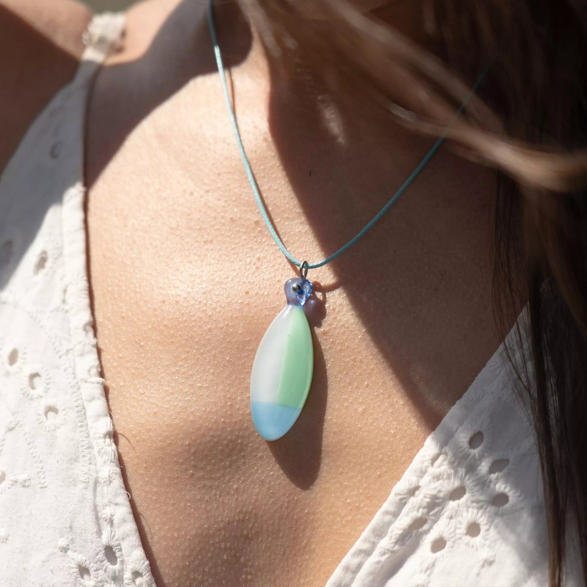Close-up of a woman wearing a Sea Breeze fused glass fish necklace with a blue cord, sunlight highlighting the green and blue tones of the pendant.