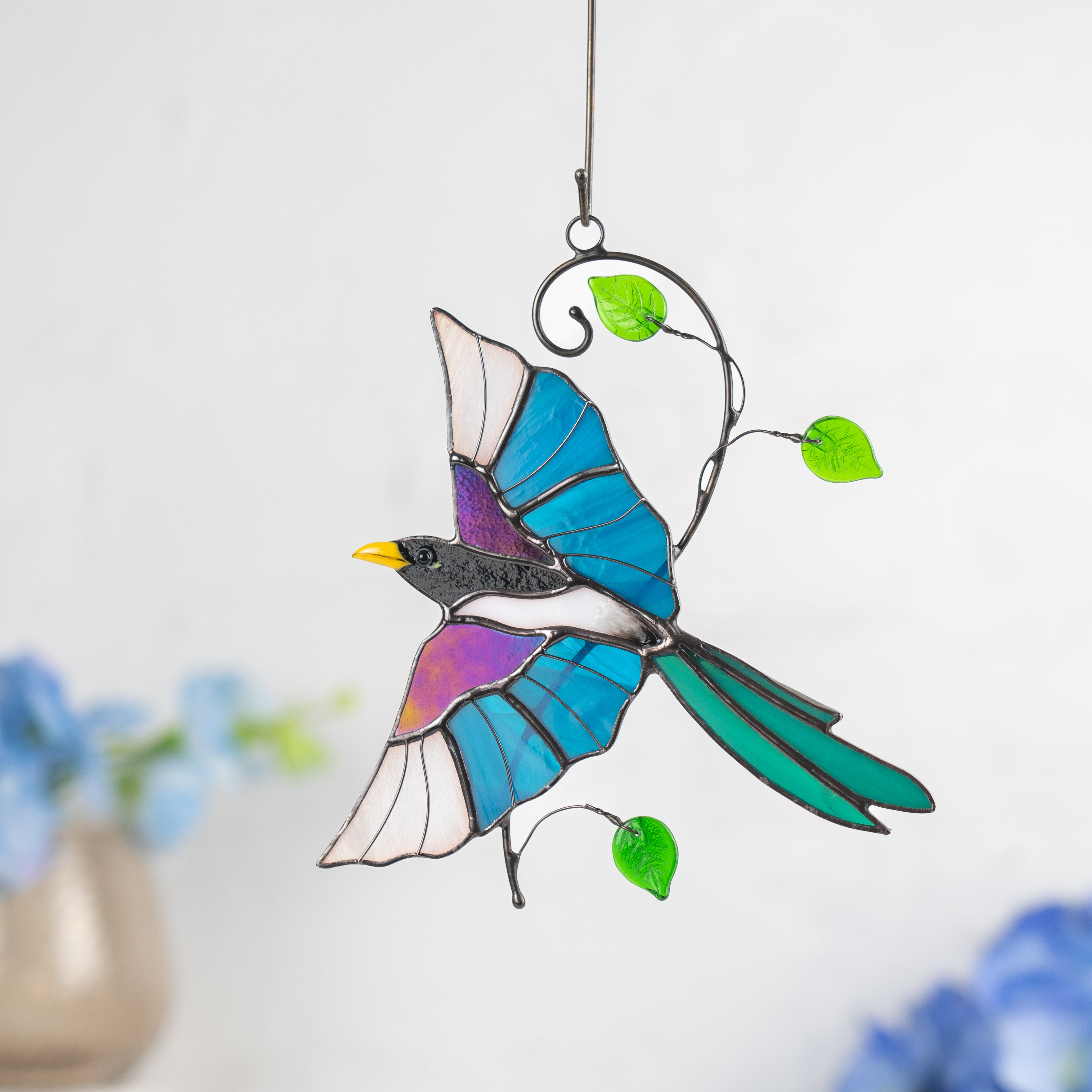 Stained glass magpie with vivid blue wings and green leaves, suspended before softly blurred blue flowers.