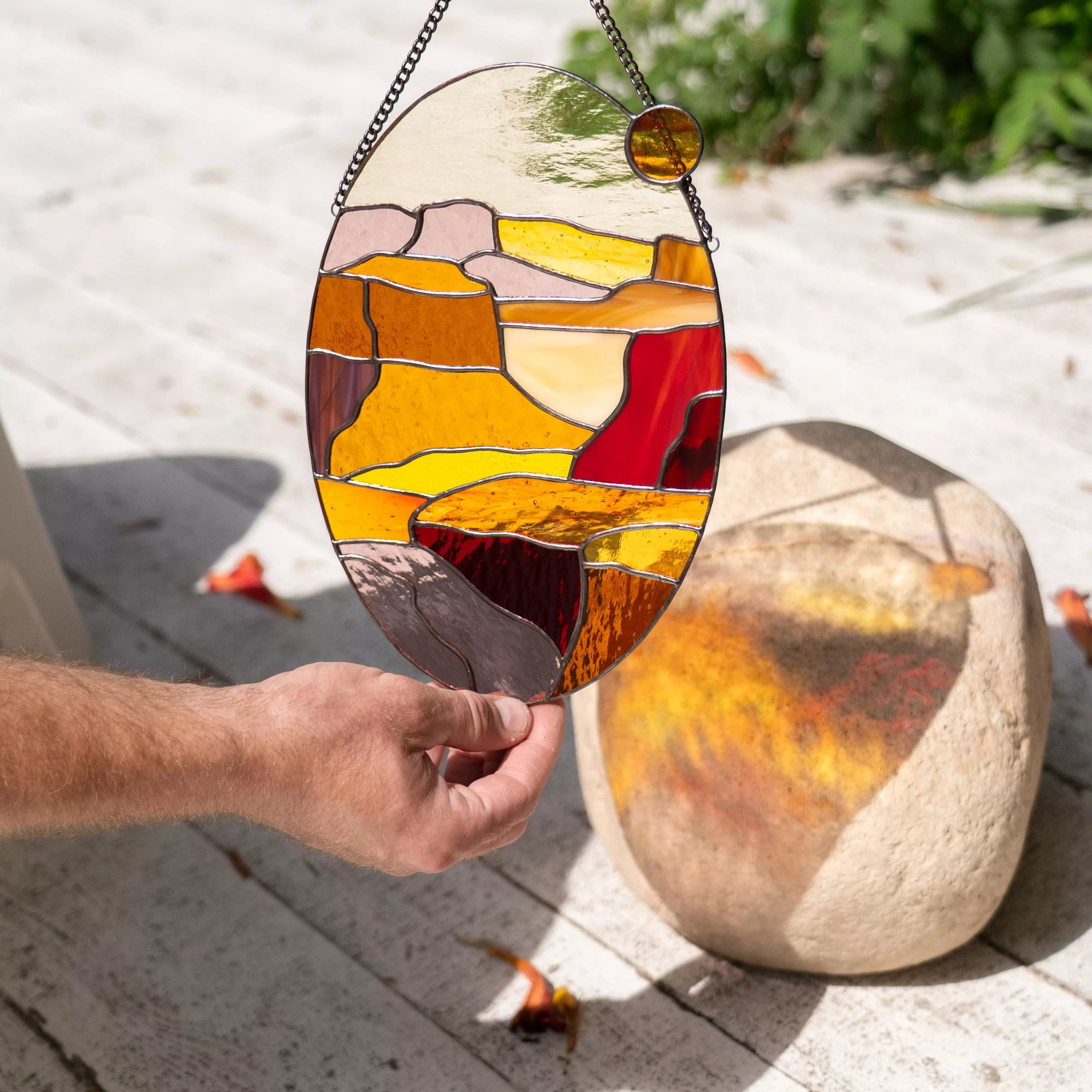 Hand holding stained glass landscape suncatcher outdoors with shadow reflection on stone.