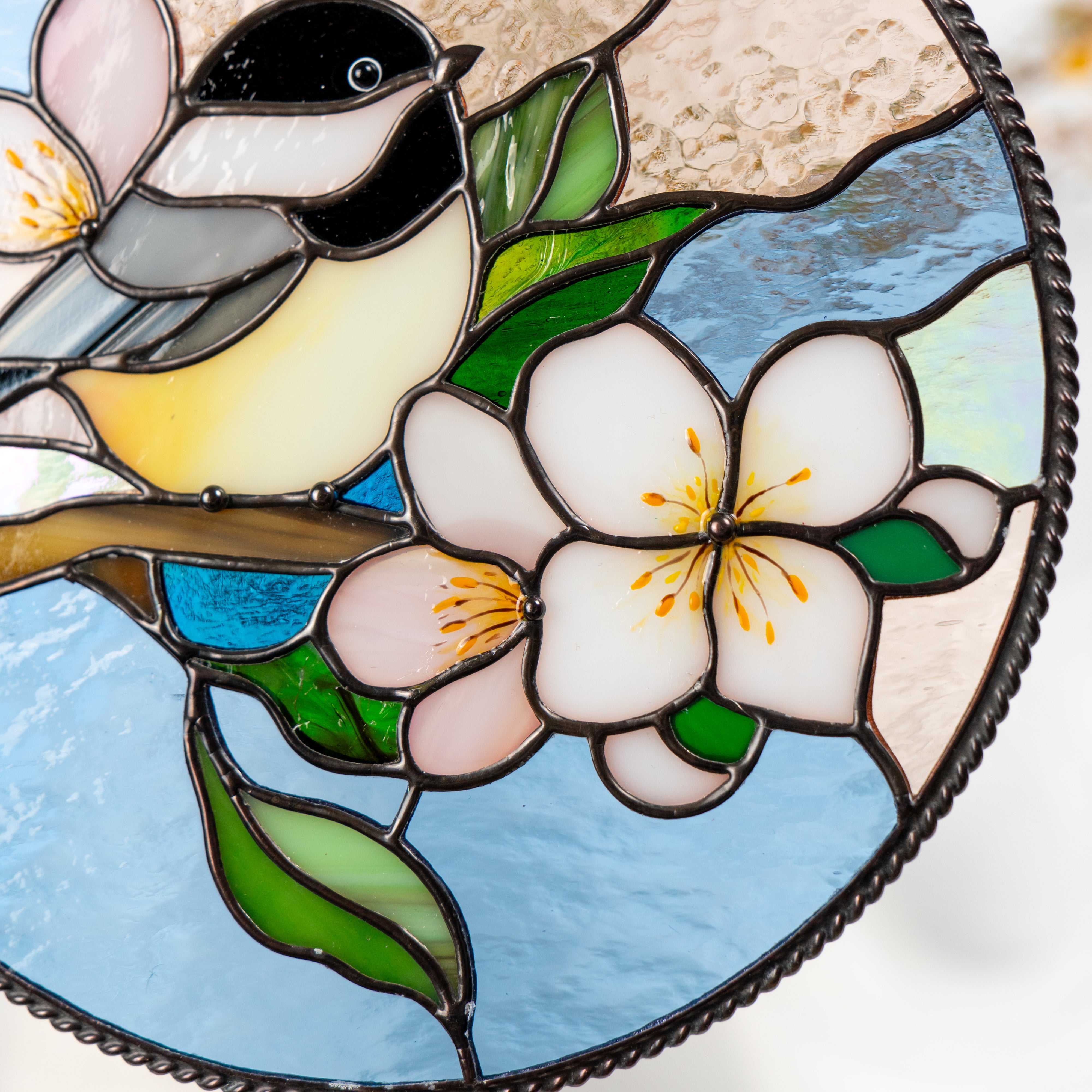 Close-up of two stained glass flowers in pink and white with yellow centers and green leaves beneath the chickadee