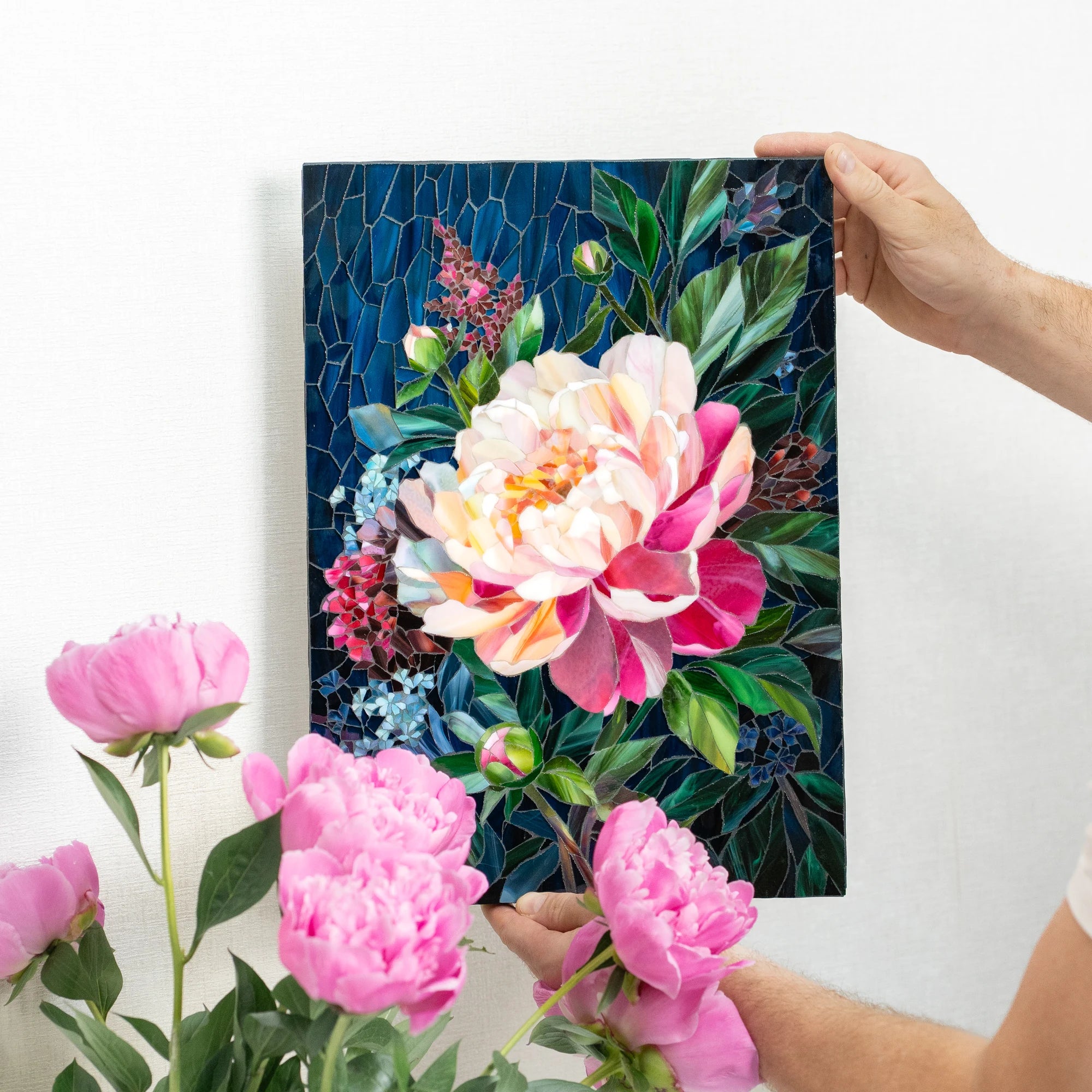 Hands holding a peony stained glass mosaic beside fresh pink peonies, showing how lifelike the hand-cut glass petals look — handmade stained glass floral wall art for the home