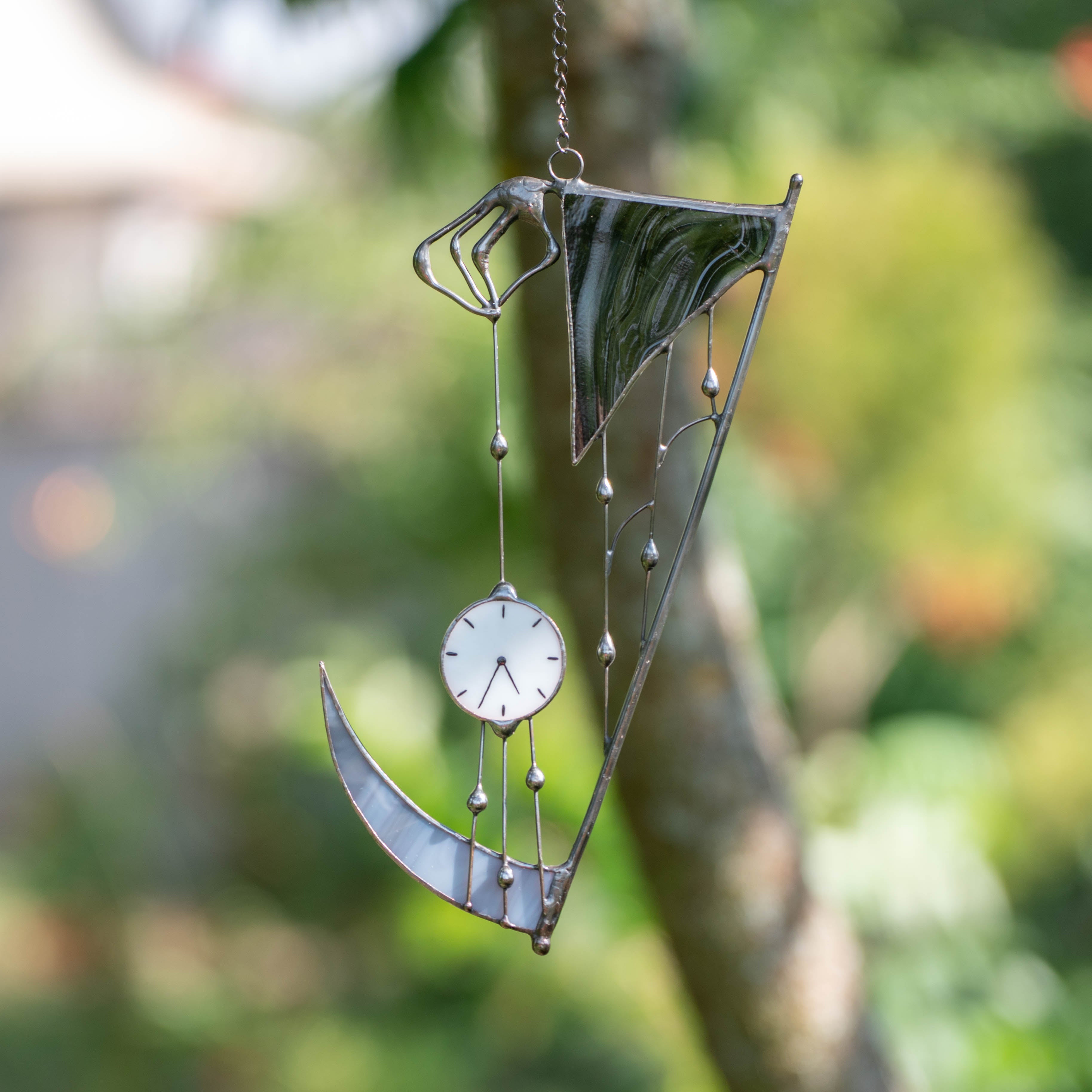 Stained glass “Hand of Death” suncatcher hanging outdoors on a tree branch, shaped like a scythe with a white clock