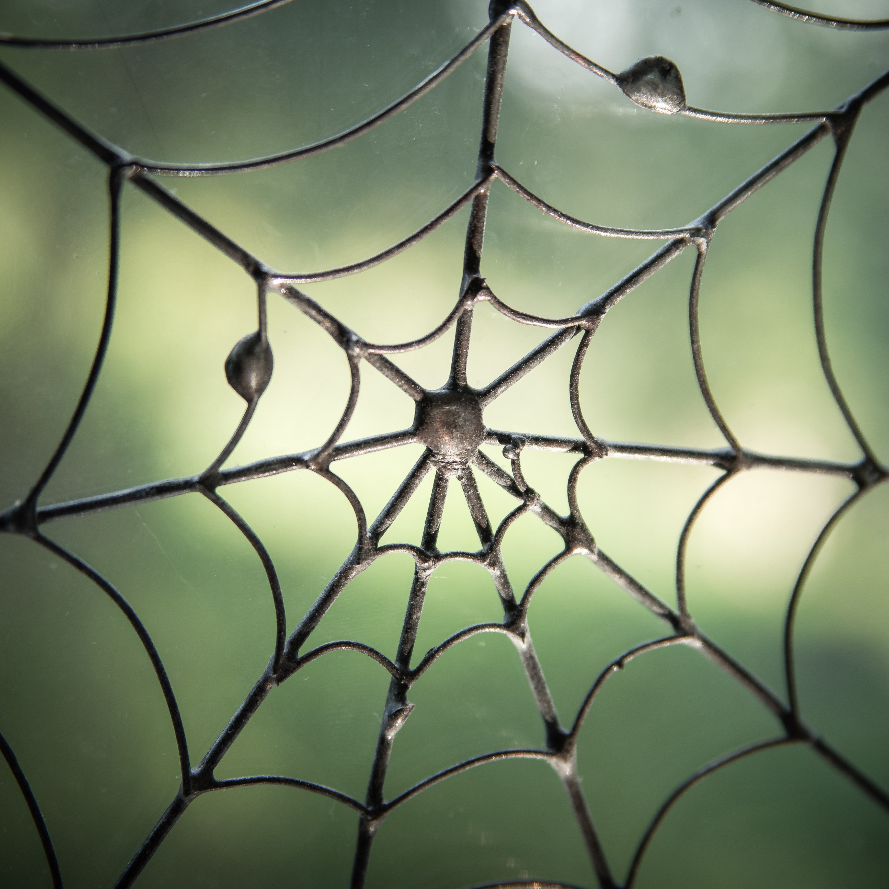 Detailed close-up of a handcrafted spider web made of black wire and glass, with textured joints, against a blurred green background.