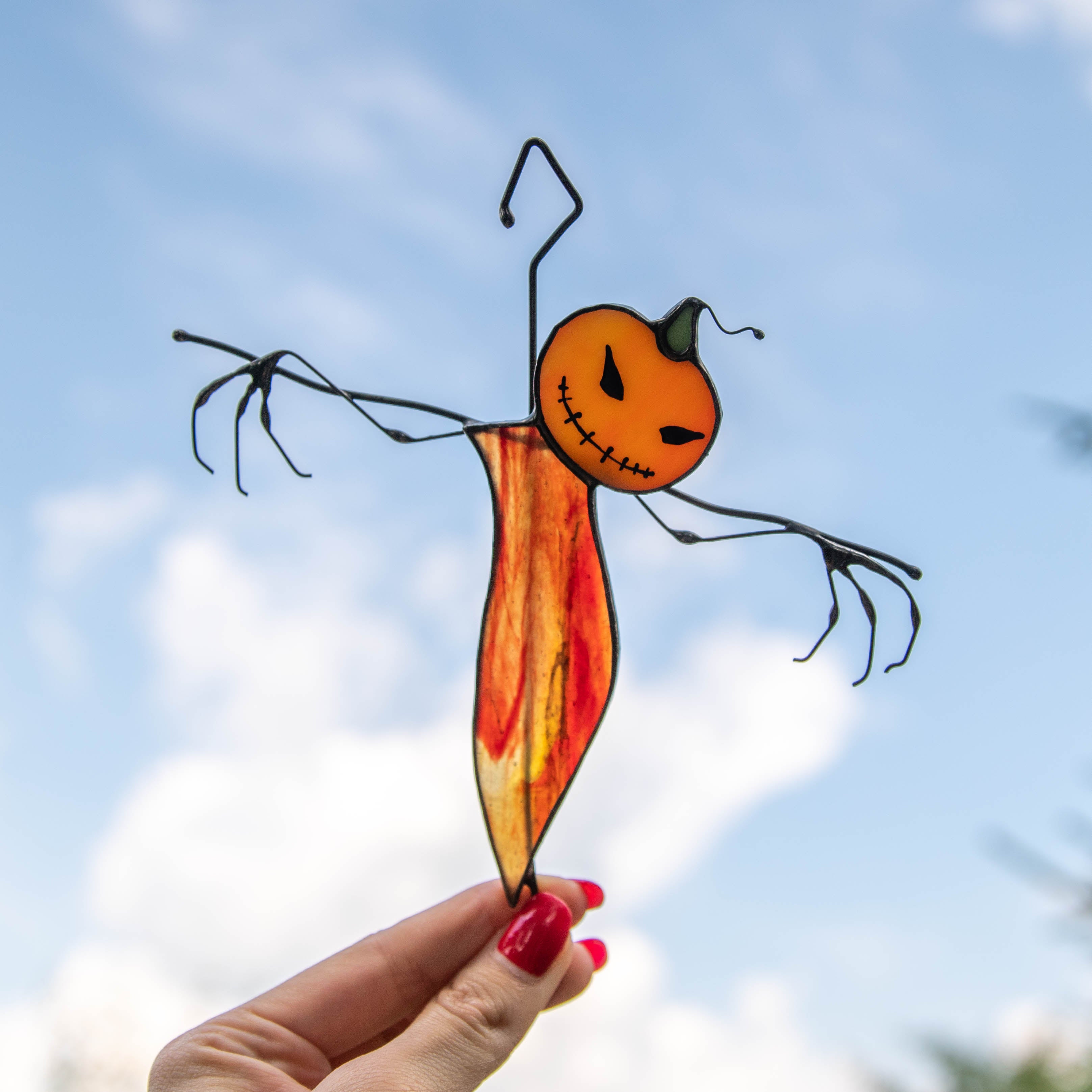 Scarecrow figure made of stained glass with wire arms and an orange head held up against the sky.