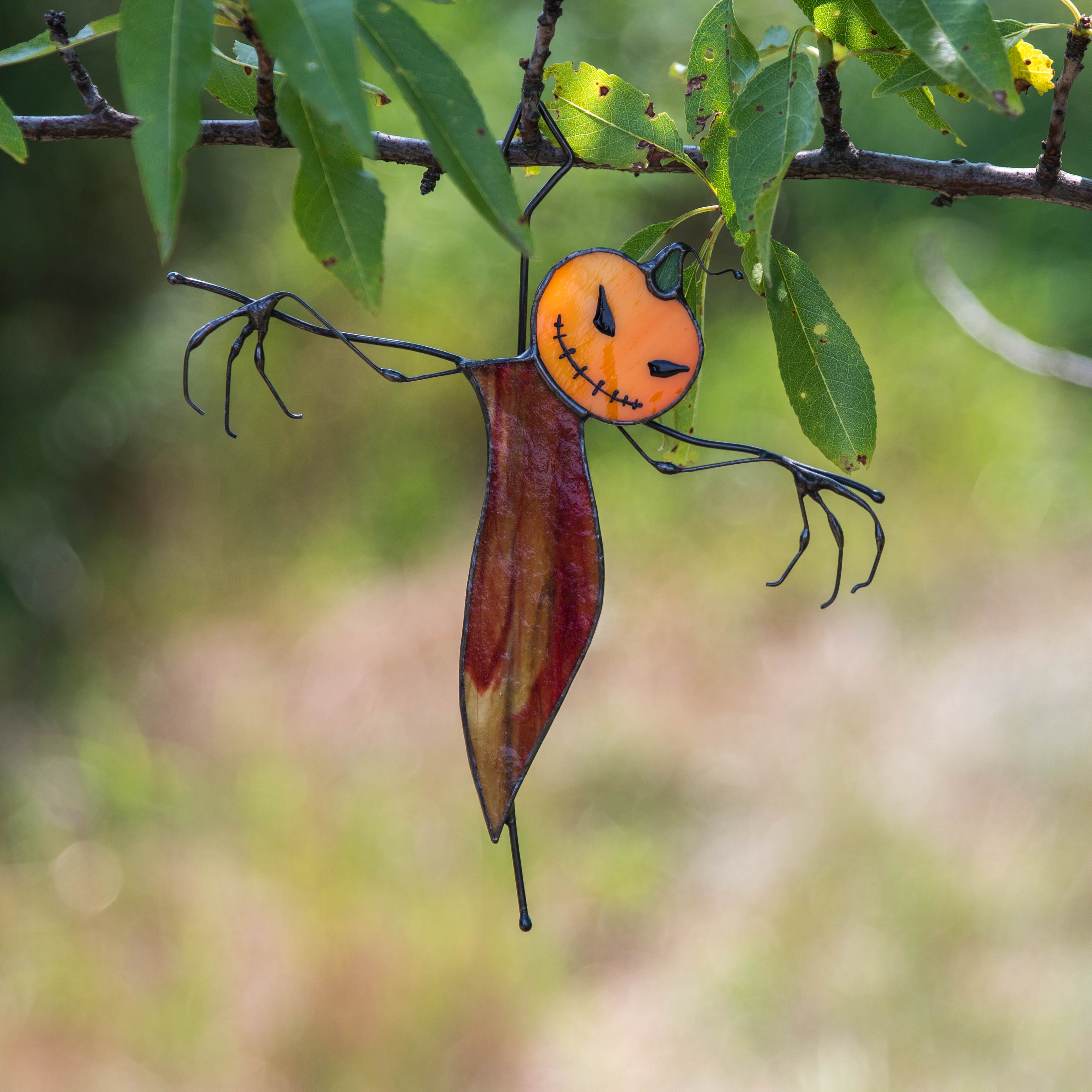Stained glass scarecrow with orange head and twisted arms hanging from a tree branch outdoors.