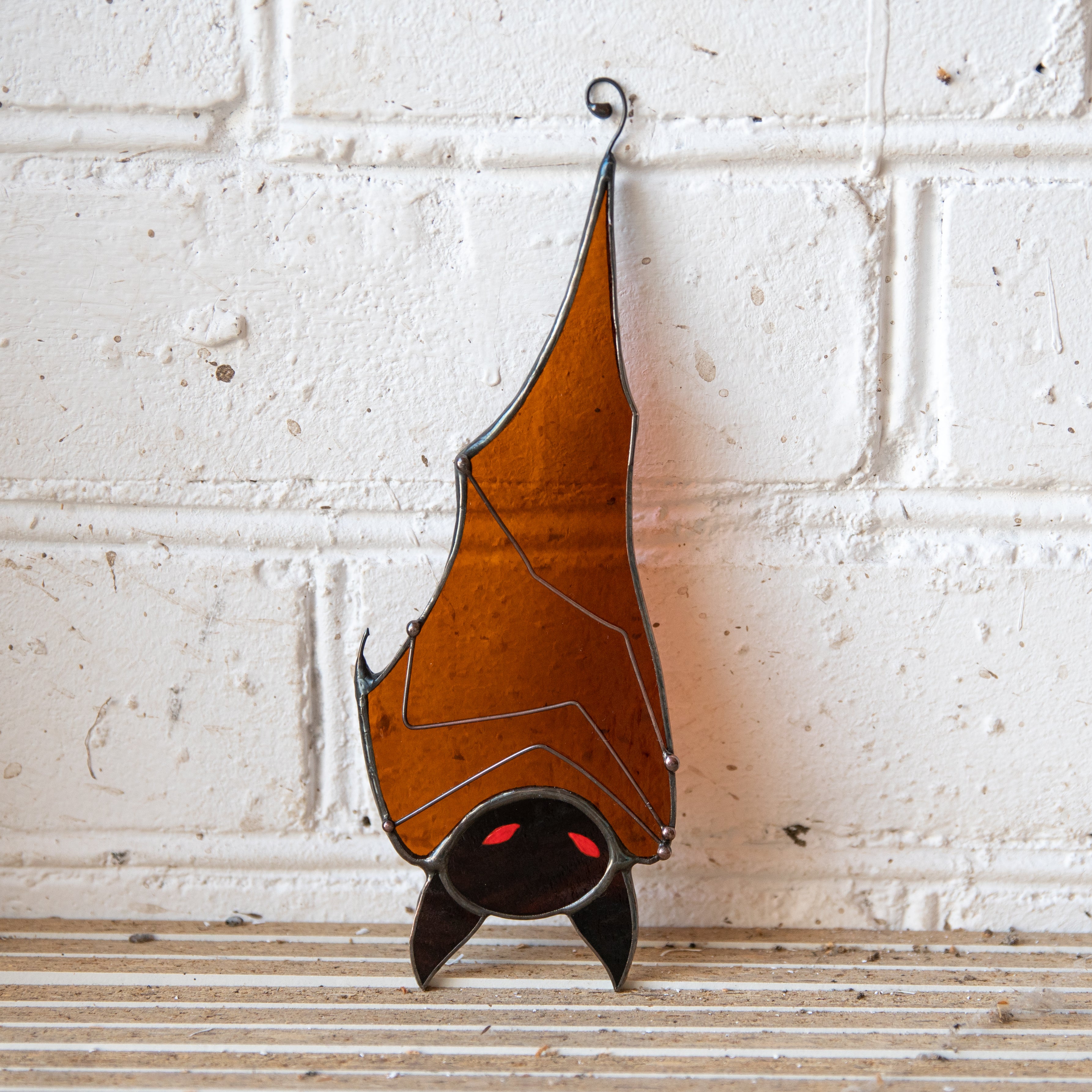 Stained glass suncatcher shaped like a bat with orange wings and glowing red eyes placed against a white brick wall.