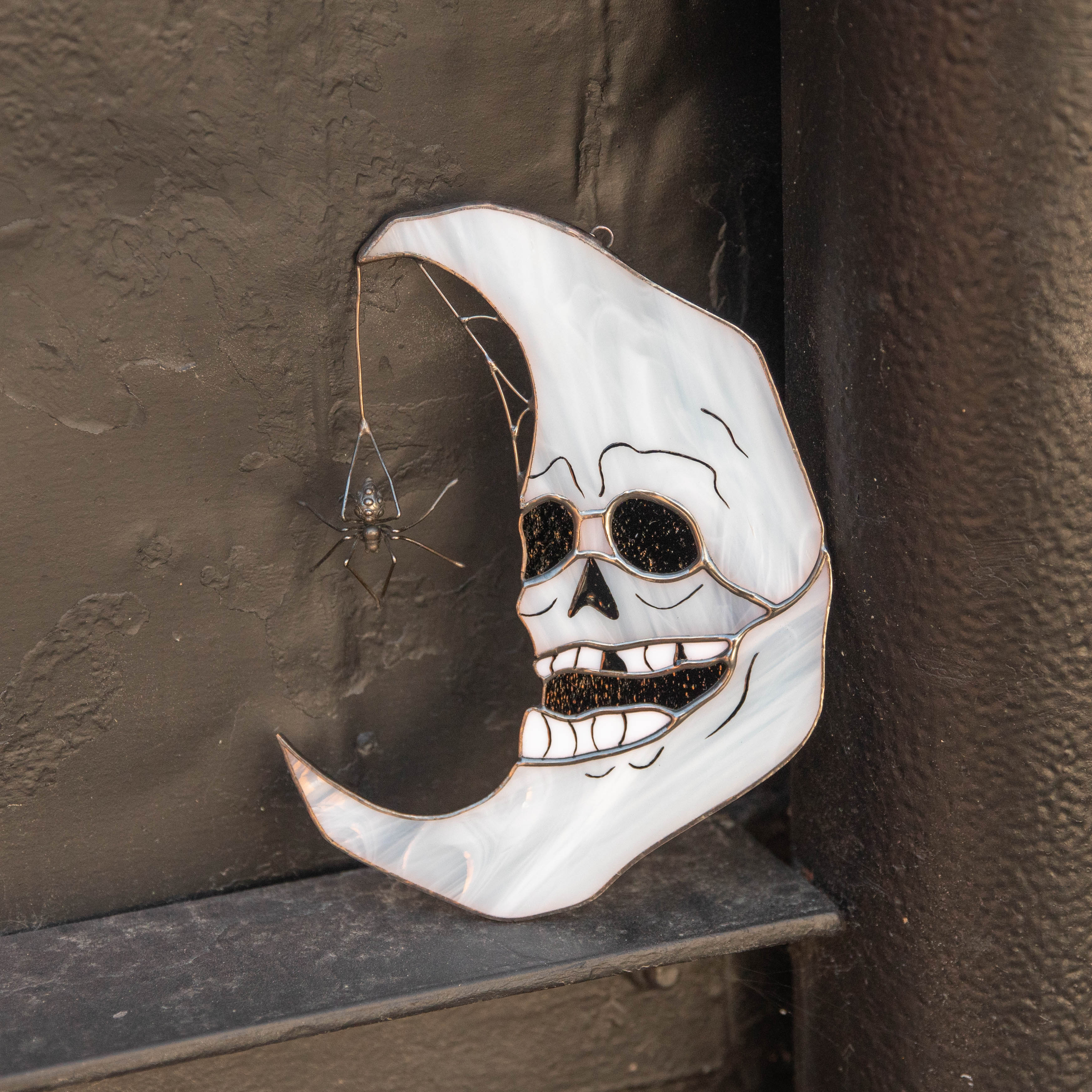 Close-up of skull moon suncatcher leaning on black wall, with spider hanging from the curved tip of the moon