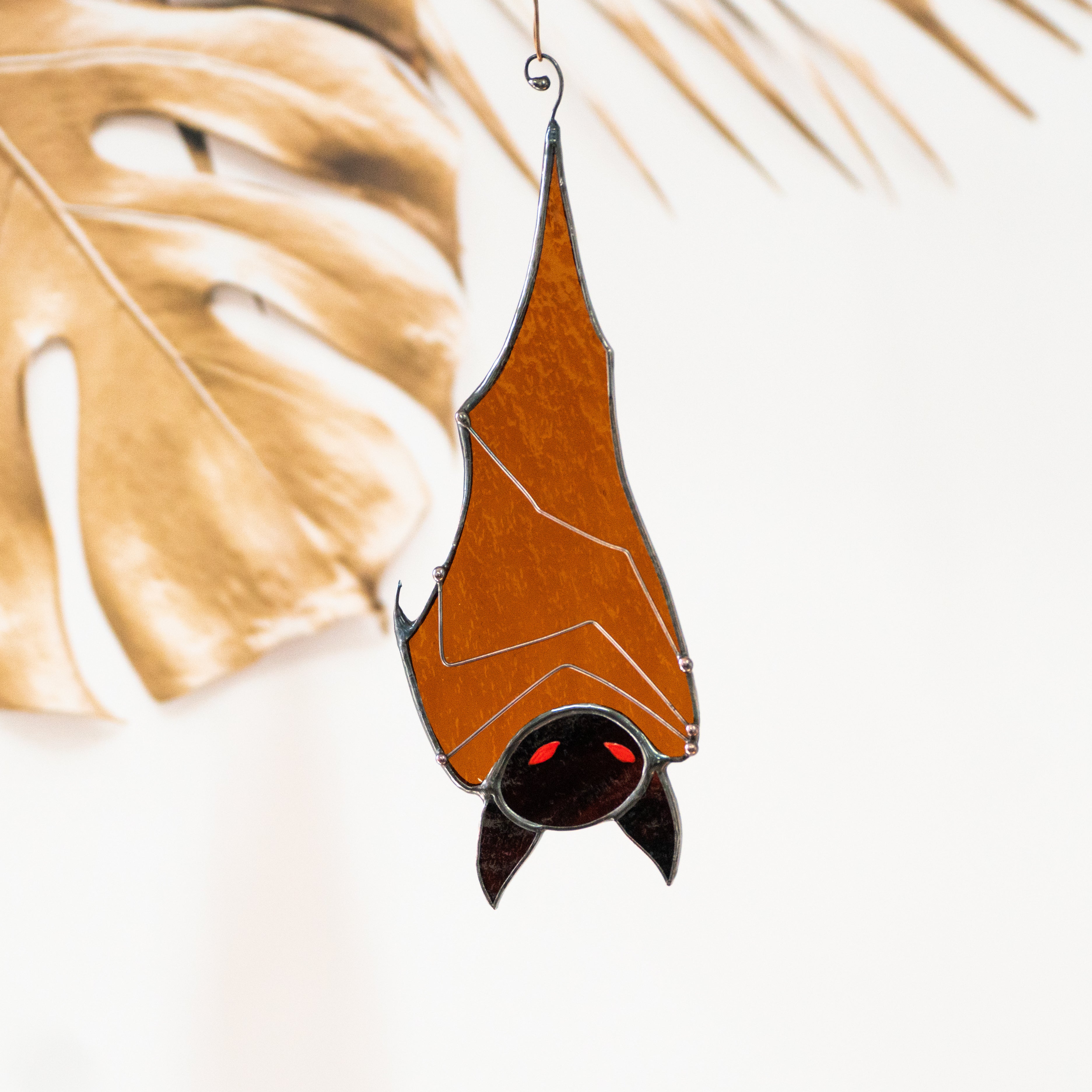 Transparent orange stained glass bat with black face and red eyes hanging in front of abstract leaf decor.