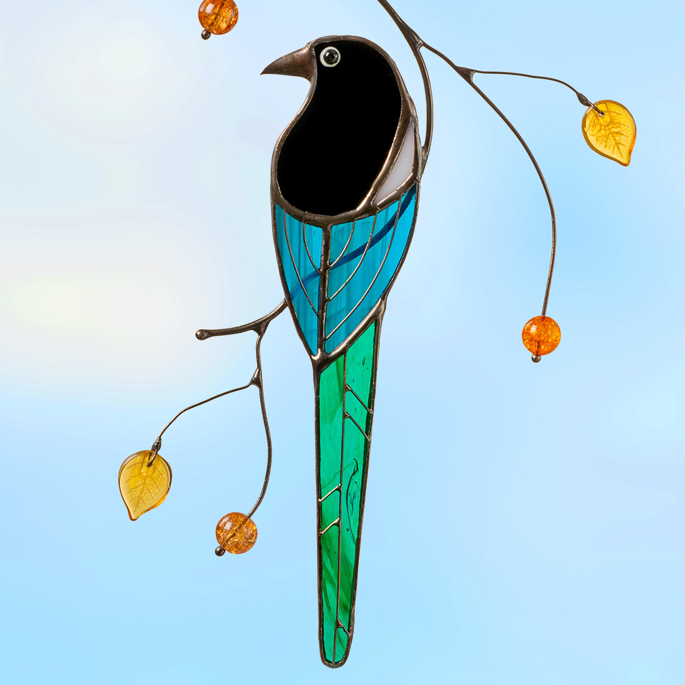 Stained glass magpie hanging outdoors against a clear blue sky, surrounded by decorative branches.