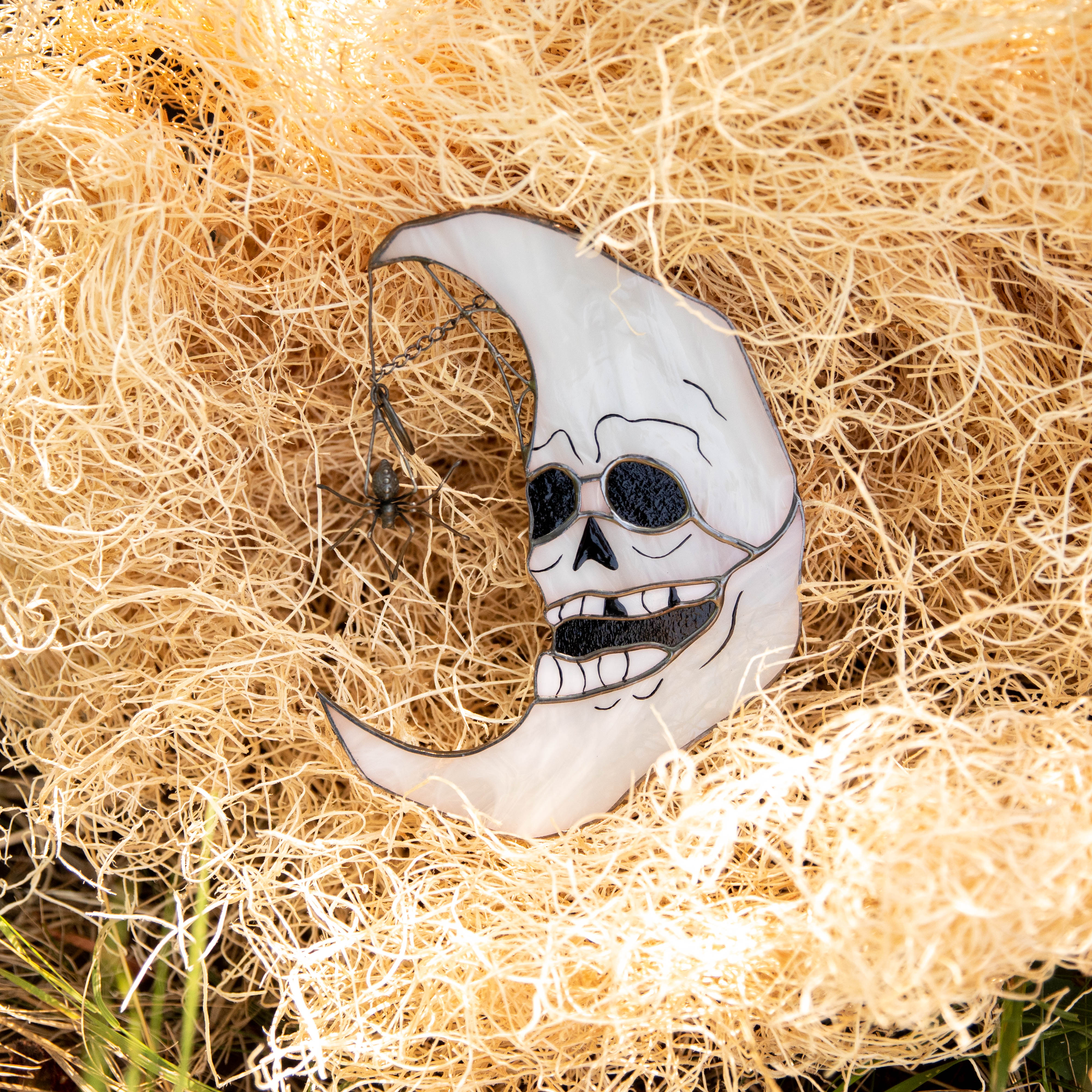 White stained glass moon shaped like a skull with a dangling black spider, resting on straw-like Halloween decorative background