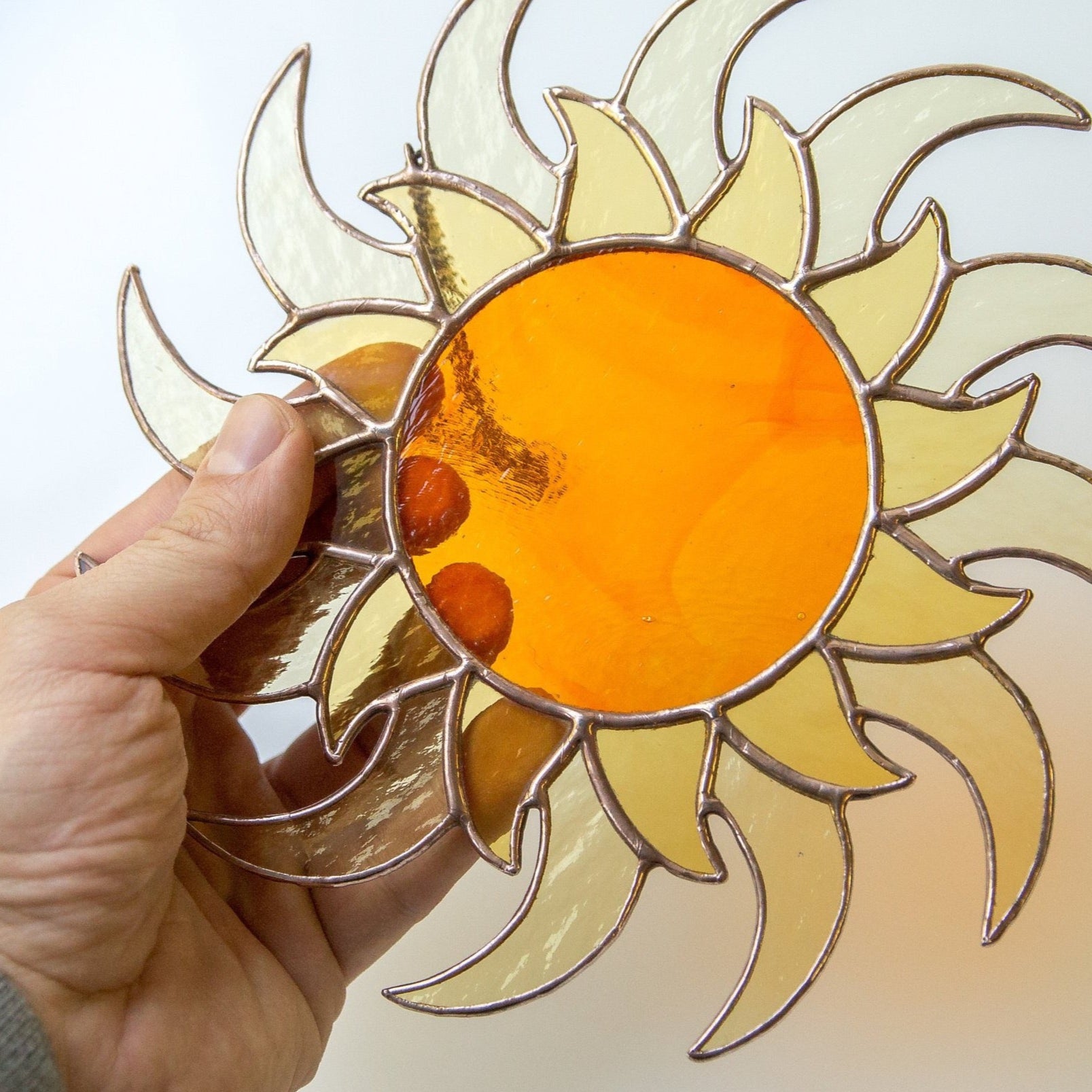 Close-up of hand holding a stained glass sun suncatcher with deep orange center and yellow-glass rays, ready to hang.