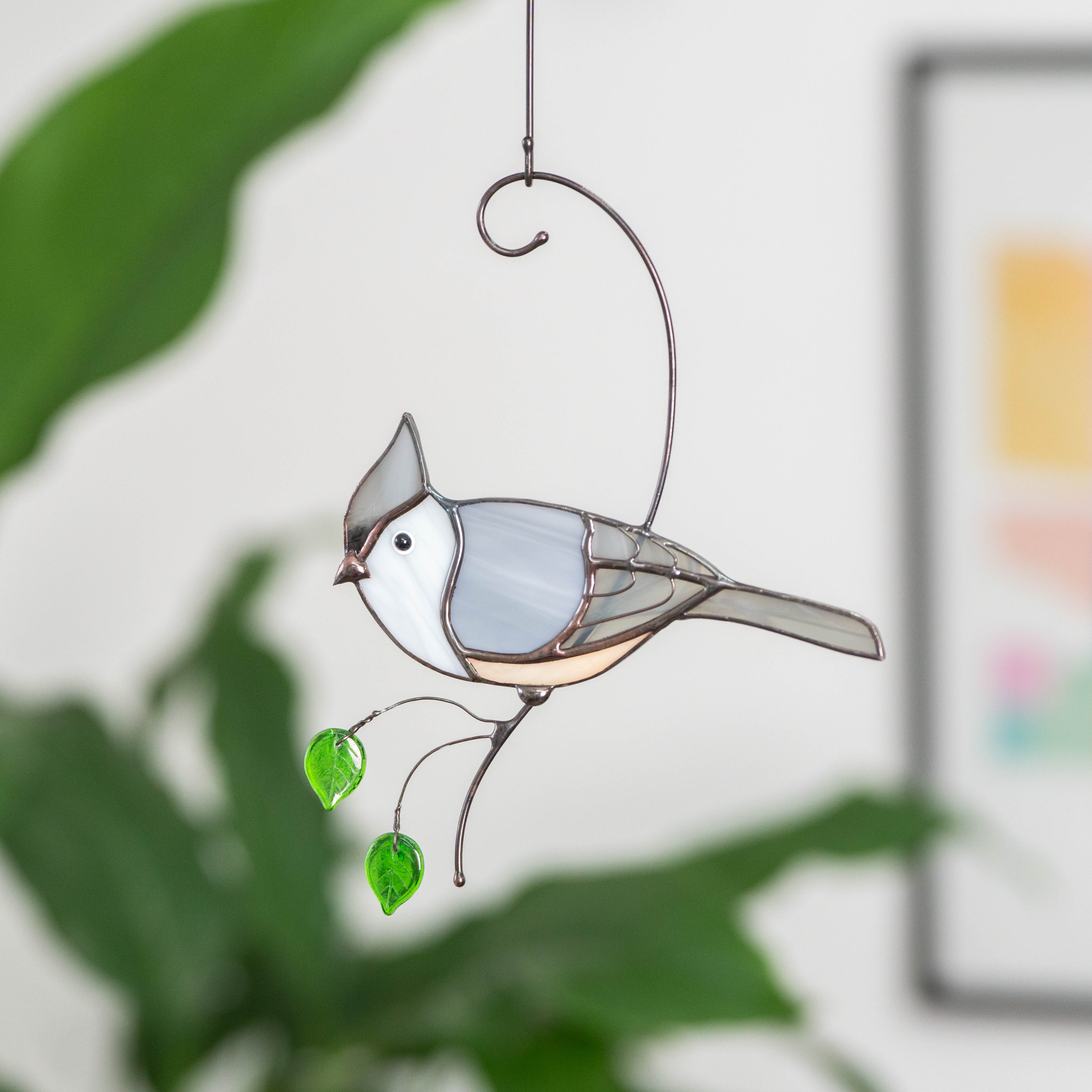 A stained glass tufted titmouse with green leaf beads, hanging against a background of green houseplants.