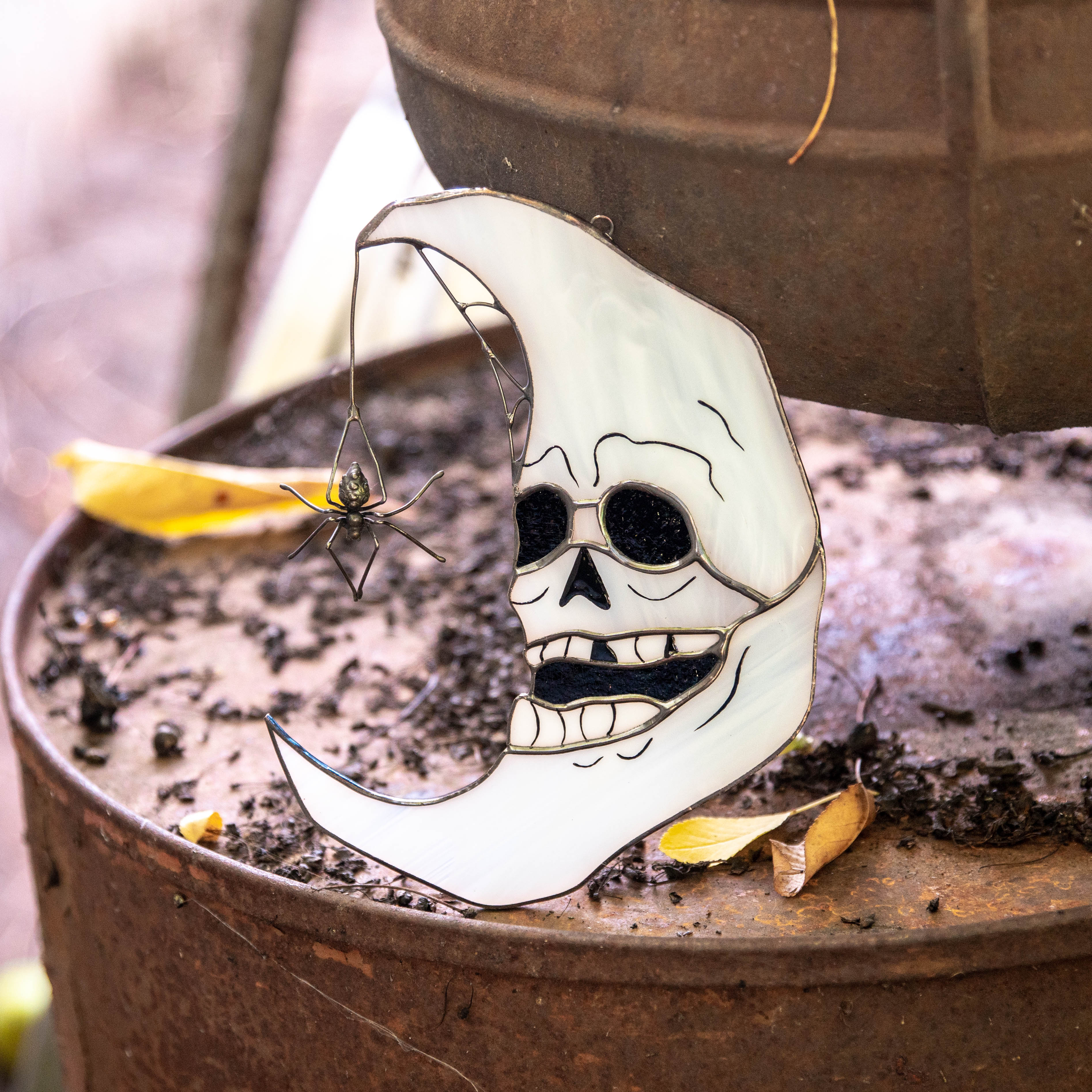Stained glass skull moon resting on rusty surface, spider dangling beside it, captured in a natural autumn scene