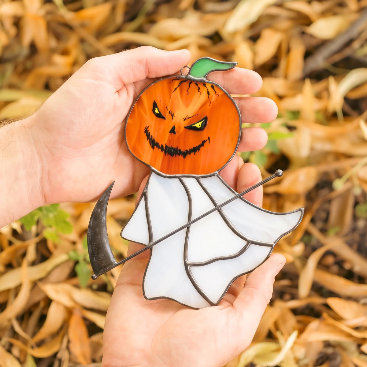 Grim reaper pumpkin head stained glass suncatcher hanging by a cord against a softly blurred green garden background.
