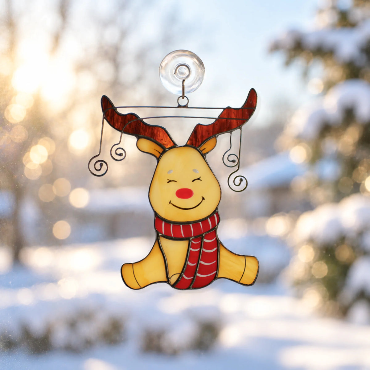 A festive stained-glass reindeer suncatcher with red antlers, a red nose, and a red-and-white striped scarf, suspended by a clear hook against a snowy outdoor window background.