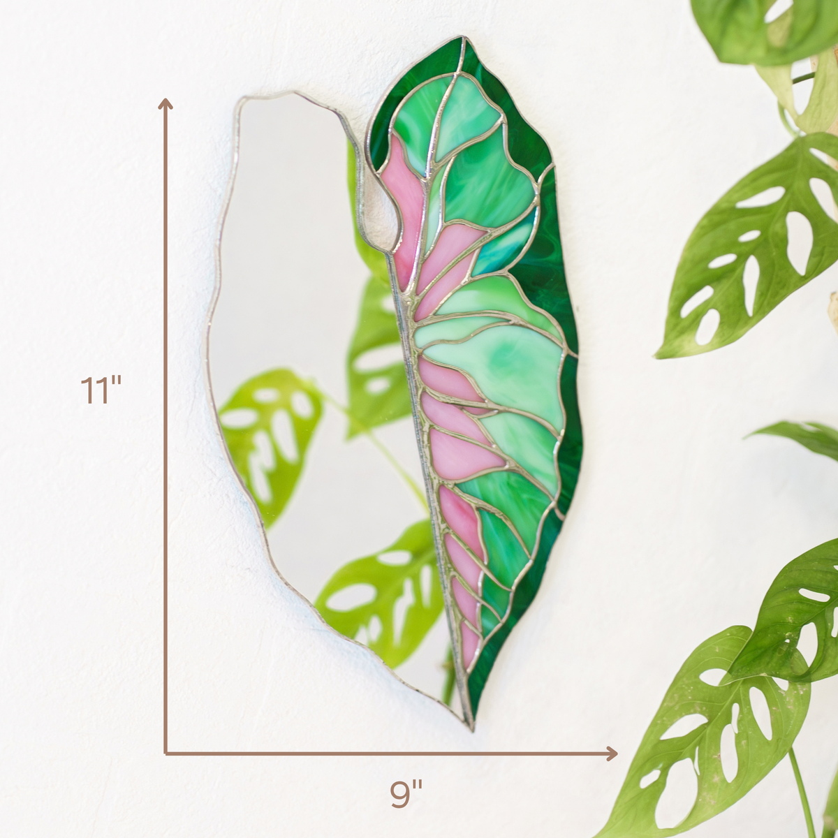 Caladium Leaf Stained Glass Mirror hanging on a white wall beside a monstera plant, measuring 11 inches tall by 9 inches wide - handmade stained glass wall decor in pink and green glass