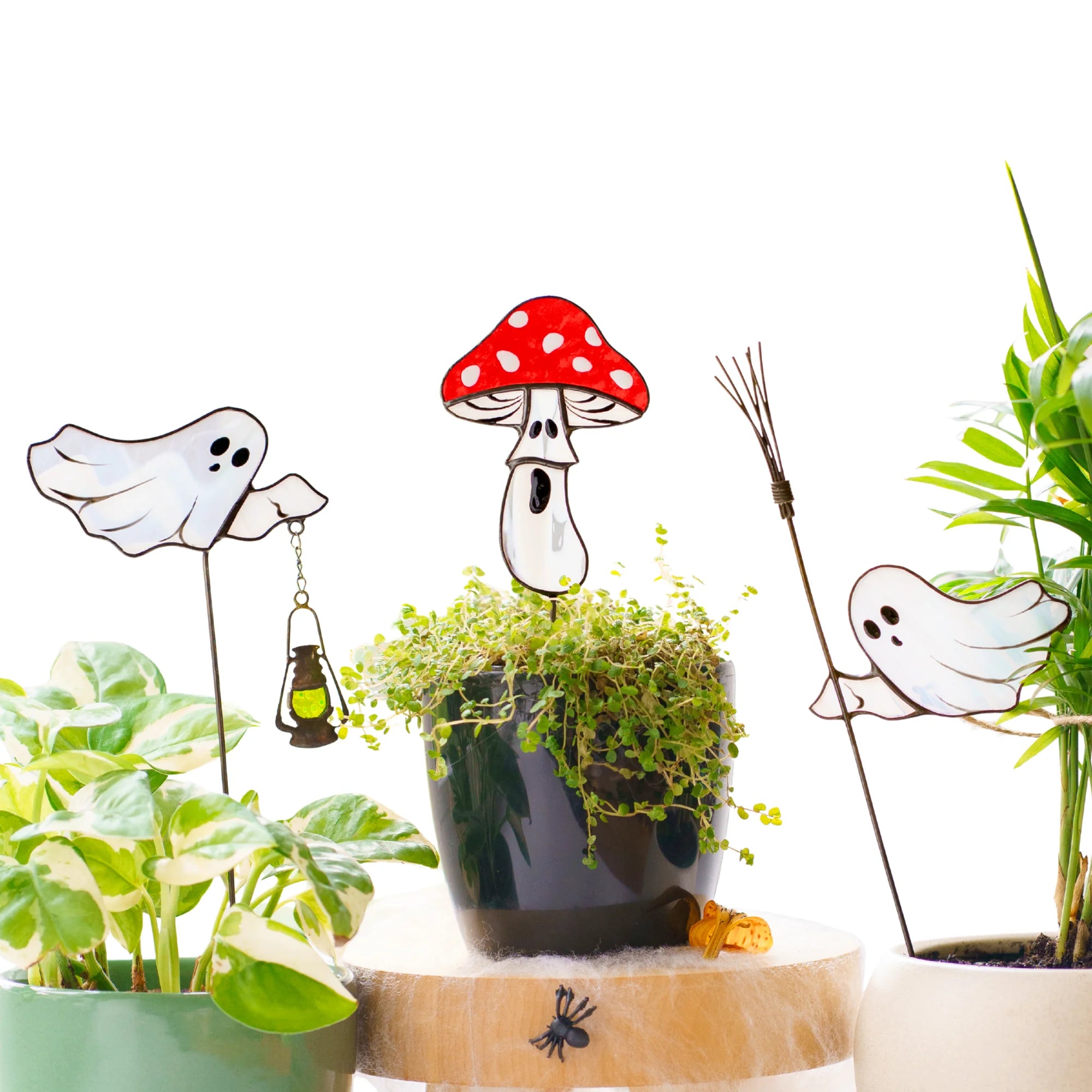 Handmade Halloween stained glass plant stakes on a white background - glass ghost with lantern, red fly agaric mushroom and ghost with broom set into potted houseplants and ivy