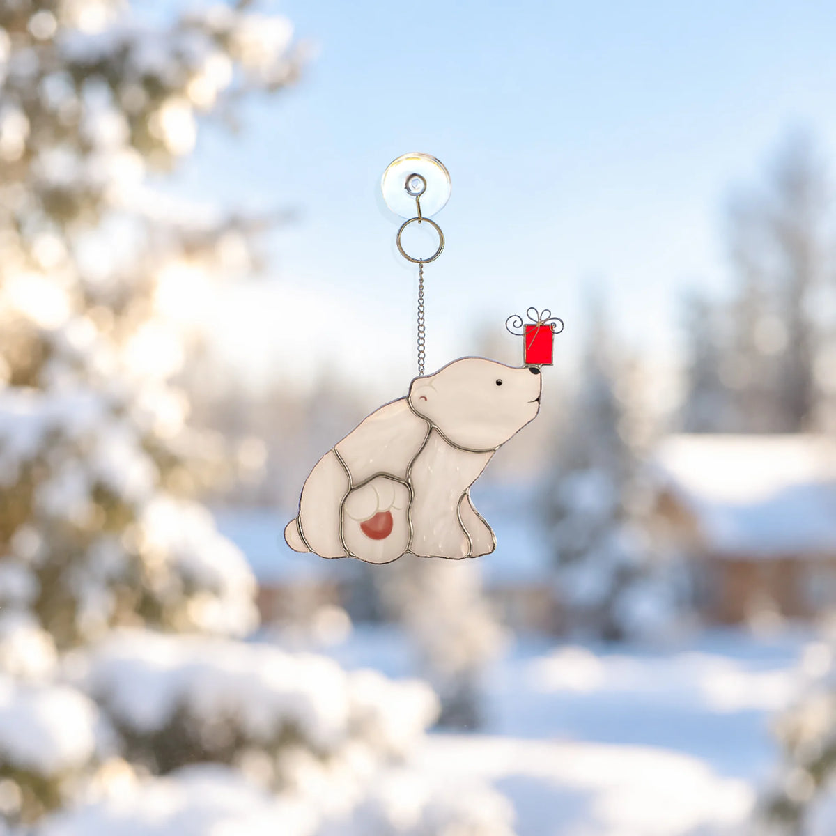 Polar bear with a Christmas gift stained glass suncatcher balancing a red gift box on its nose, hanging by a suction cup on a window against a sunny snowy forest - handmade Christmas window decor