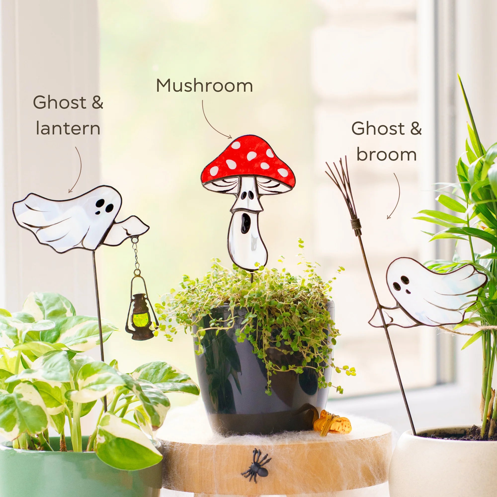 Three Halloween-themed garden stakes displayed on potted plants in front of a window. The stakes include ghost and lantern, mushroom, and ghost and broom designs with handwritten labels pointing to them.