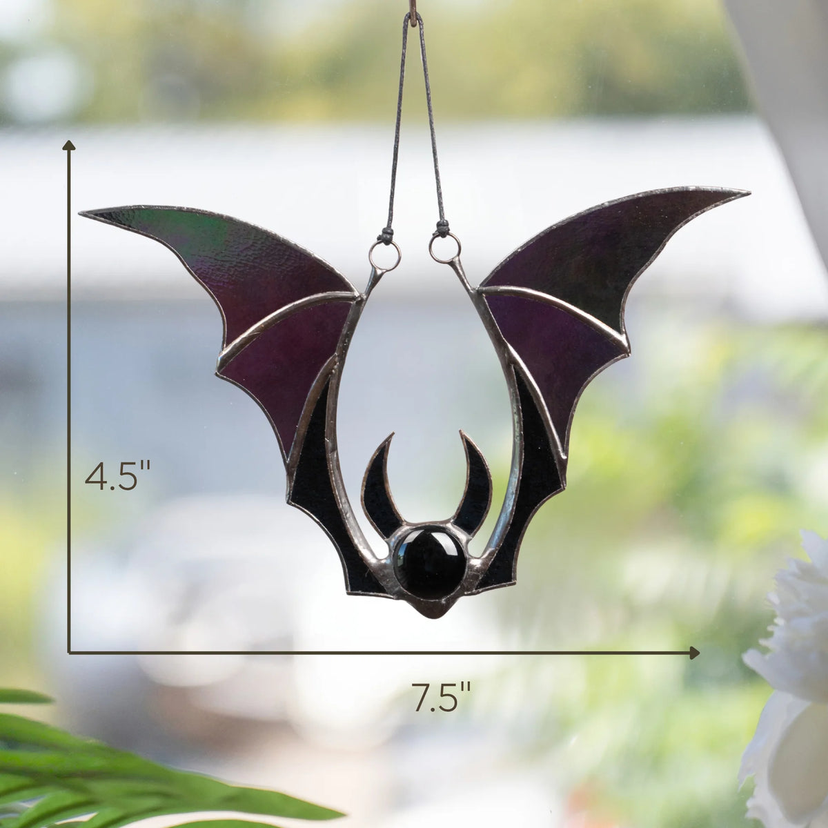 Size guide for the handmade iridescent bat stained glass suncatcher - 7.5 inches wide and 4.5 inches tall, hanging on a black cord in a bright window