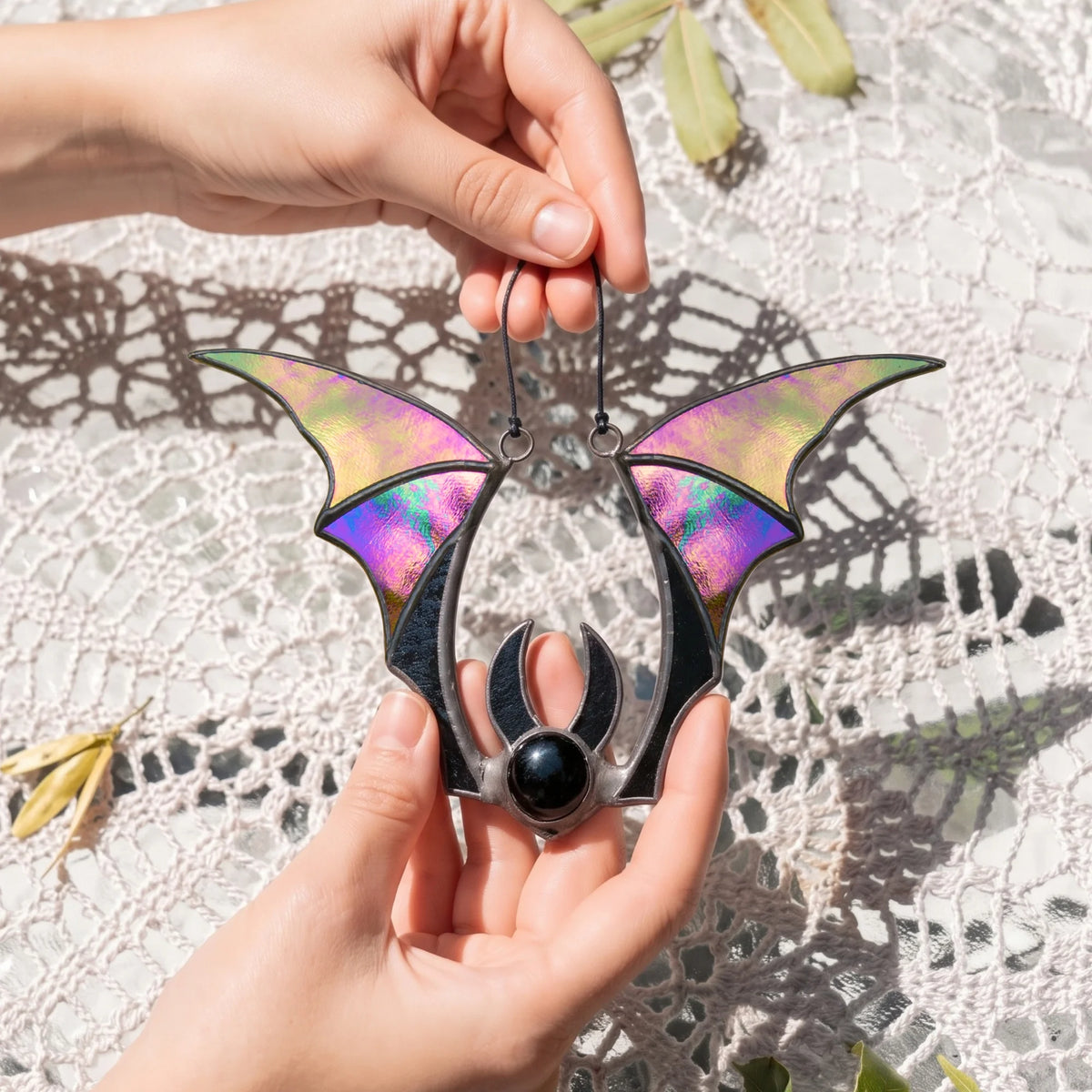 Handmade iridescent bat stained glass suncatcher resting in two open palms over a lace tablecloth, the wings glowing pink, gold and green in the sun