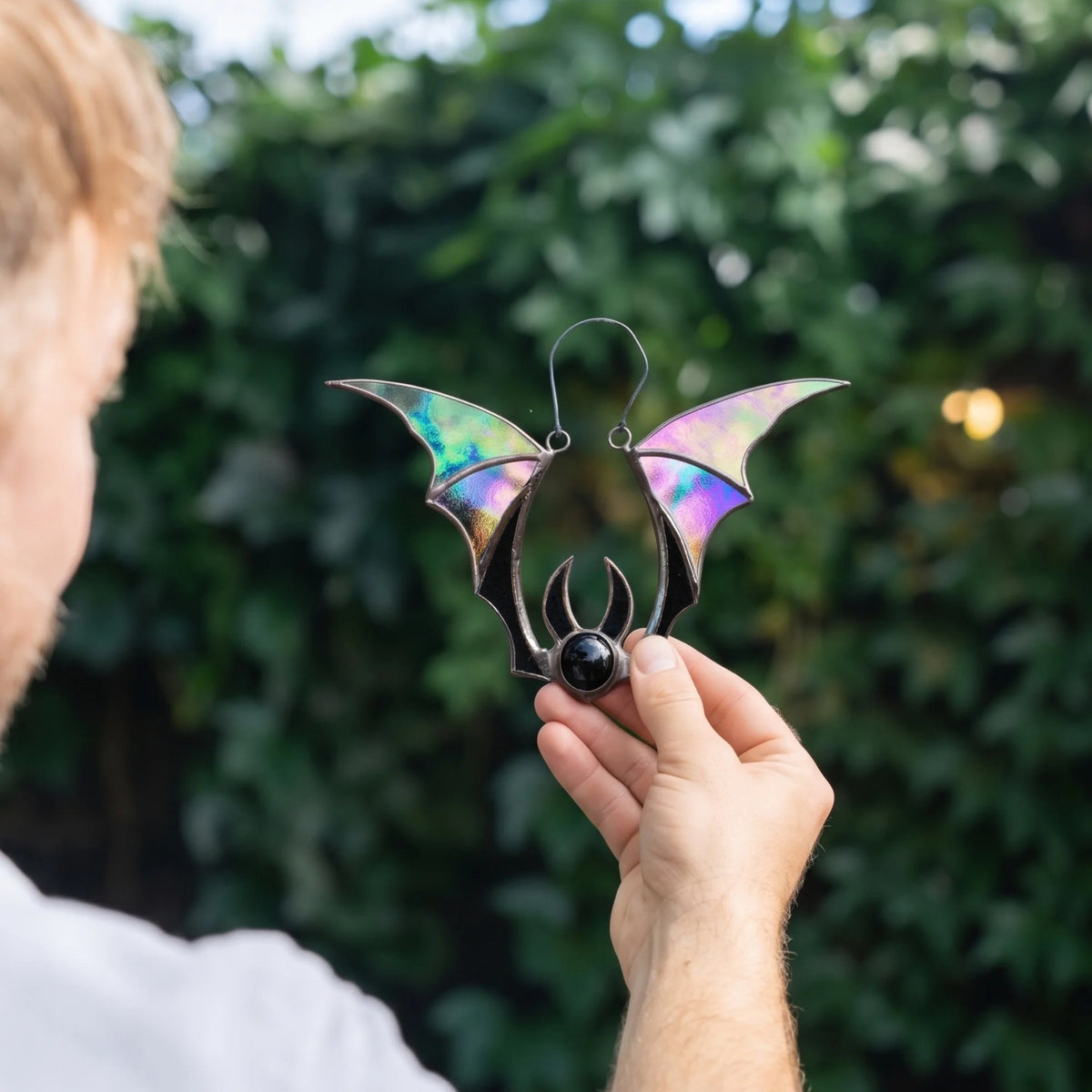Man holding the iridescent bat Halloween stained glass suncatcher up against a green hedge, the wings shifting through lilac, mint and gold as the light moves