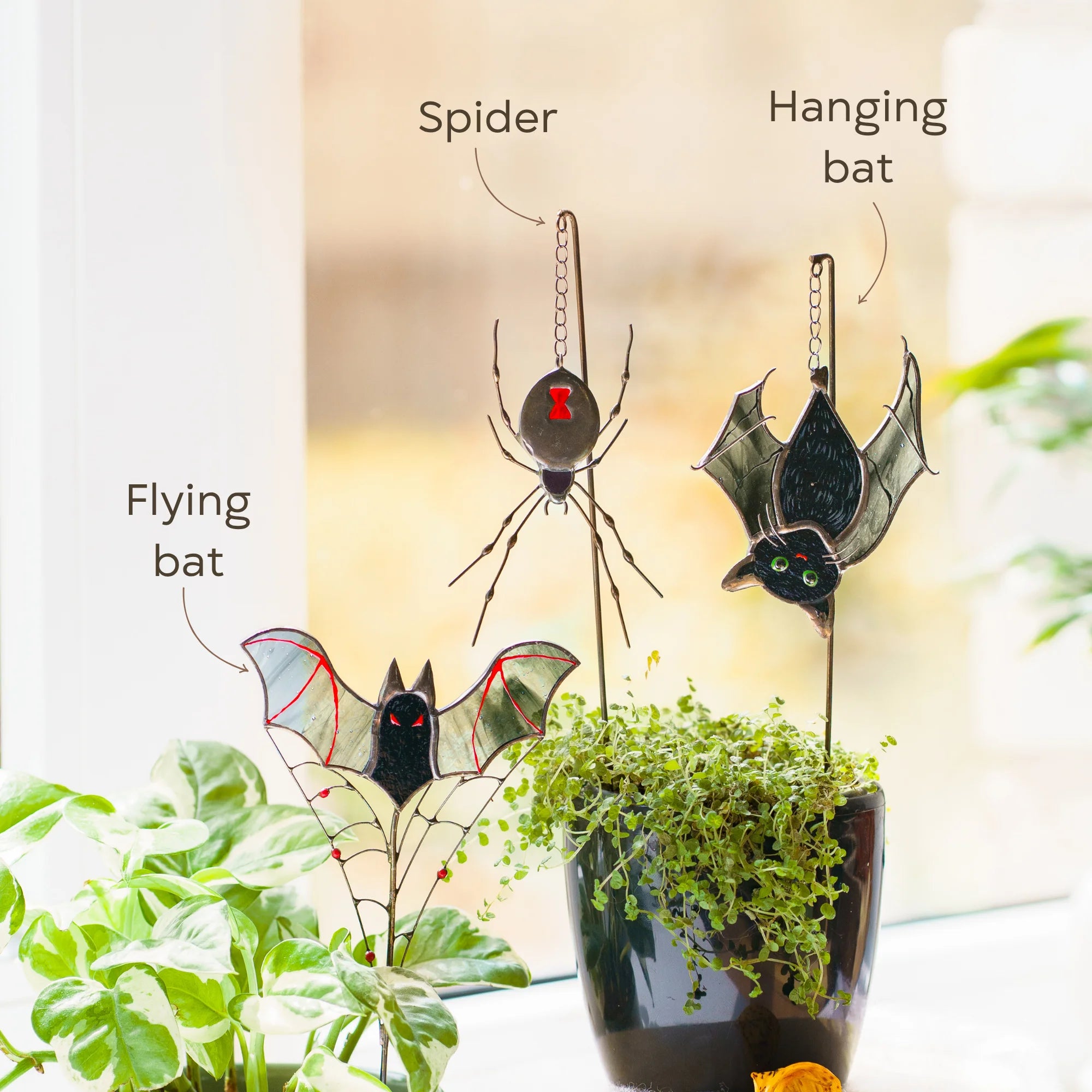 Three Halloween-themed metal garden stakes displayed on a windowsill next to potted plants. The set includes a spider stake, a hanging bat stake, and a flying bat stake, all featuring stained-glass style wings and bodies.