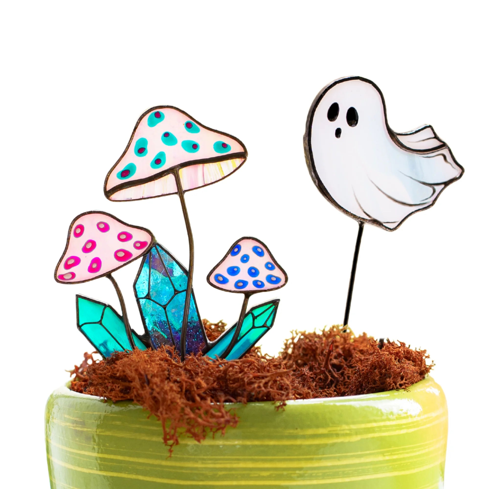 A set of stained glass plant stakes shaped like mushrooms and a ghost, arranged in brown moss inside a light green ceramic pot against a white background.