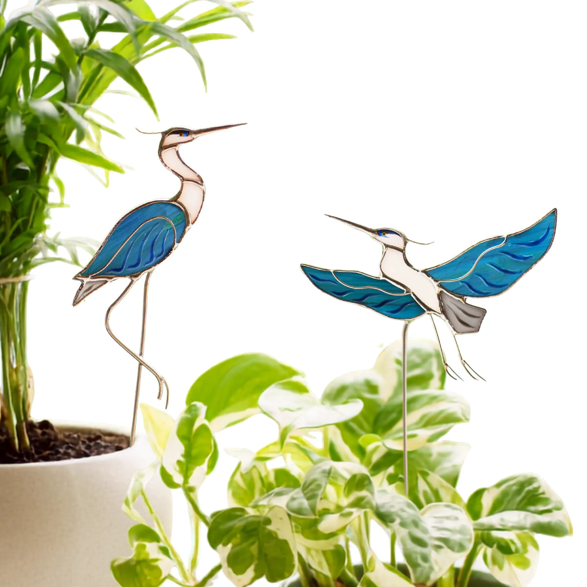 Set of two stained glass heron plant stakes - one sitting, one flying - in blue and white glass, displayed among a bamboo palm and pothos, handmade stained glass decor for plant lovers
