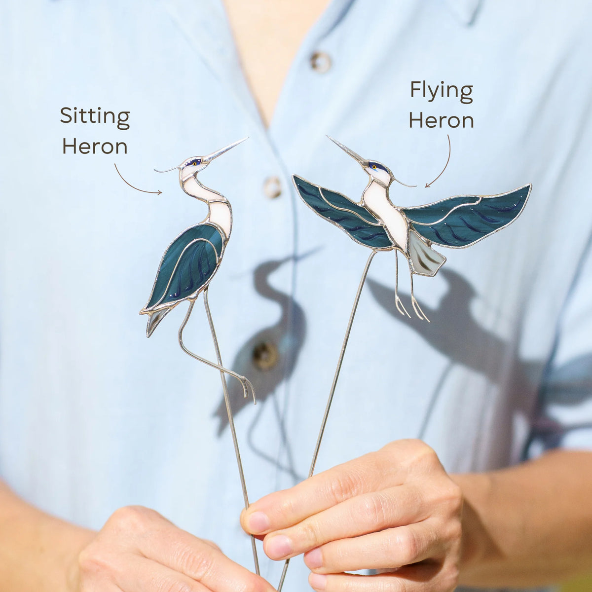 Hand holding two stained glass heron plant stakes labeled Sitting Heron and Flying Heron, blue and white glass birds casting a heron-shaped shadow in sunlight