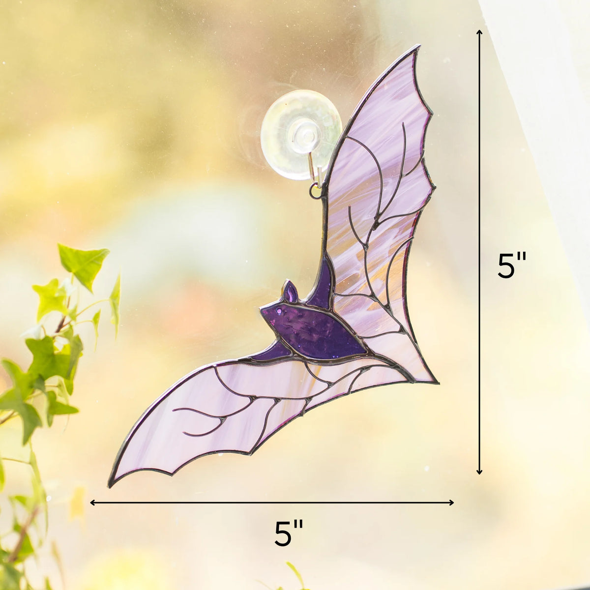 Size guide for the handmade purple bat stained glass suncatcher - 5 inches tall and 5 inches wide, hanging on a suction hook on a sunny window beside an ivy plant