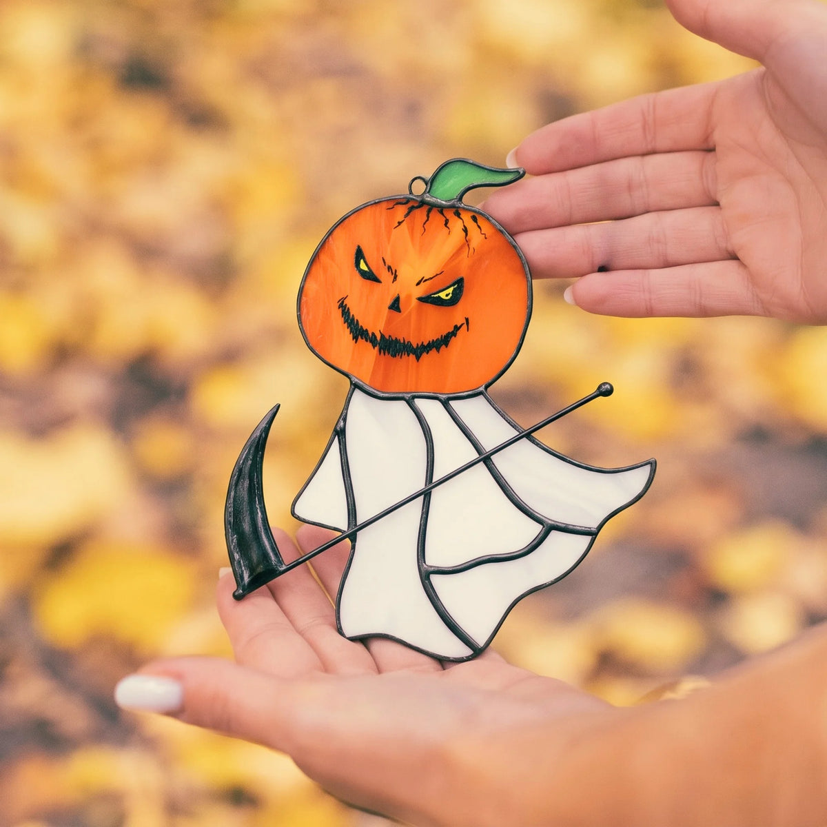 Hand holding the grim reaper pumpkin head stained glass suncatcher up close against a blurred golden autumn leaf backdrop.