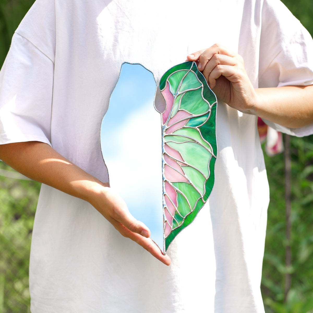 Person in a white t-shirt holding a handmade Caladium Leaf Stained Glass Mirror, the mirror half reflecting blue sky next to pink, mint and emerald glass - a unique housewarming gift