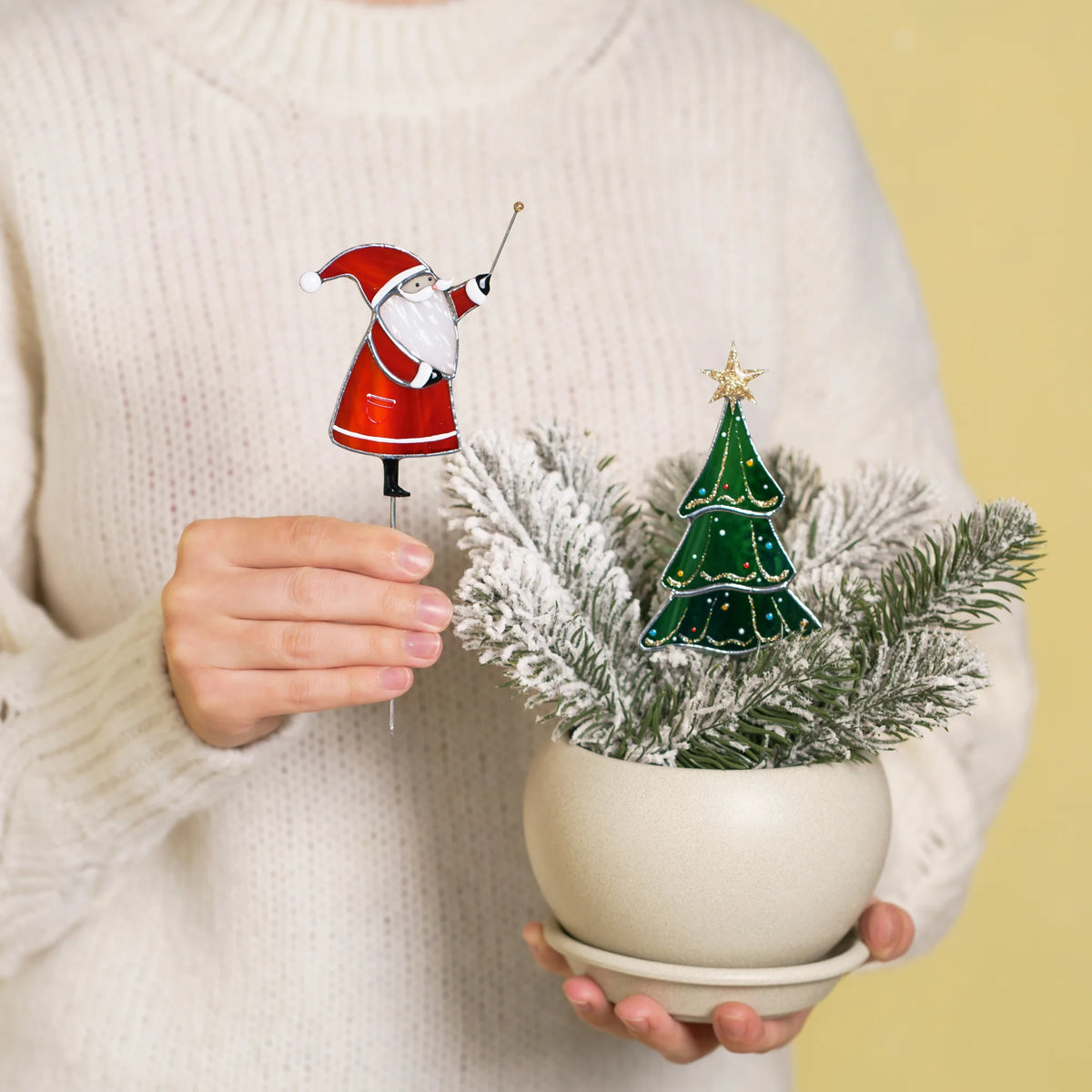 Woman in a cream sweater holding the Santa plant stake beside a white pot with frosted fir and the Christmas tree stake - handmade stained glass plant stakes and a festive holiday gift