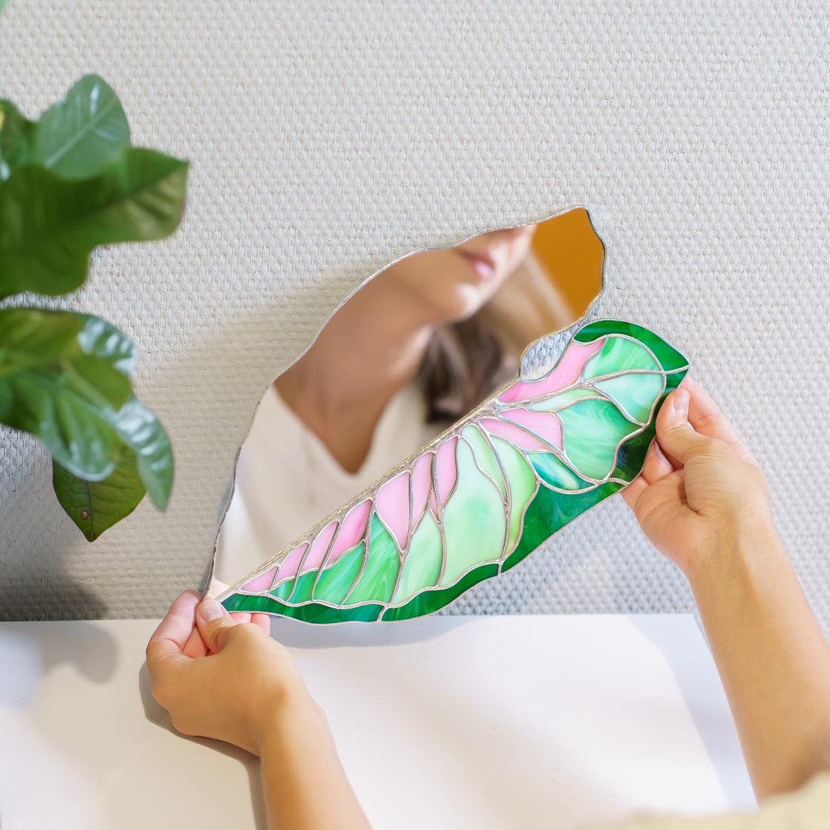 Caladium Leaf Stained Glass Mirror held at a white table, showing a woman's reflection in the mirror half beside pink and green stained glass veins - handmade mirror stained glass decor