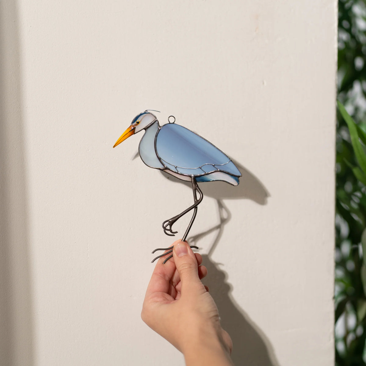 Hand holding a handmade stained glass great blue heron against a sunlit wall, its long-legged shadow stretching behind it - bird suncatcher for bright windows and plant corners