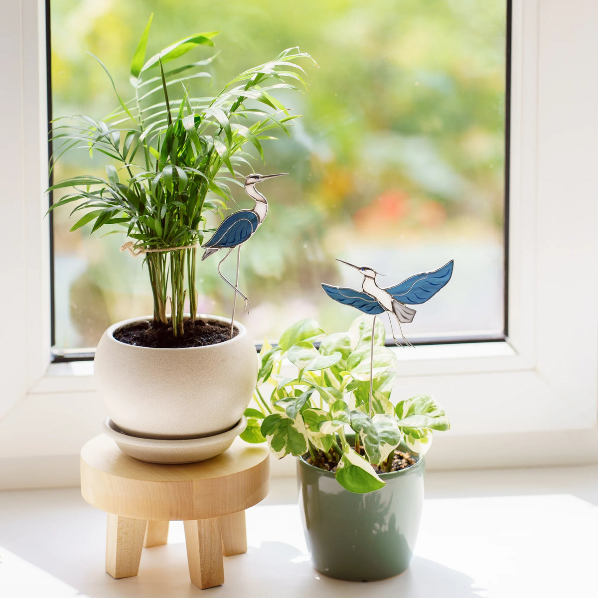Close-up of stained glass heron plant stakes - sitting heron beside a palm and flying heron among variegated pothos - on a sunny windowsill, handmade stained glass decor